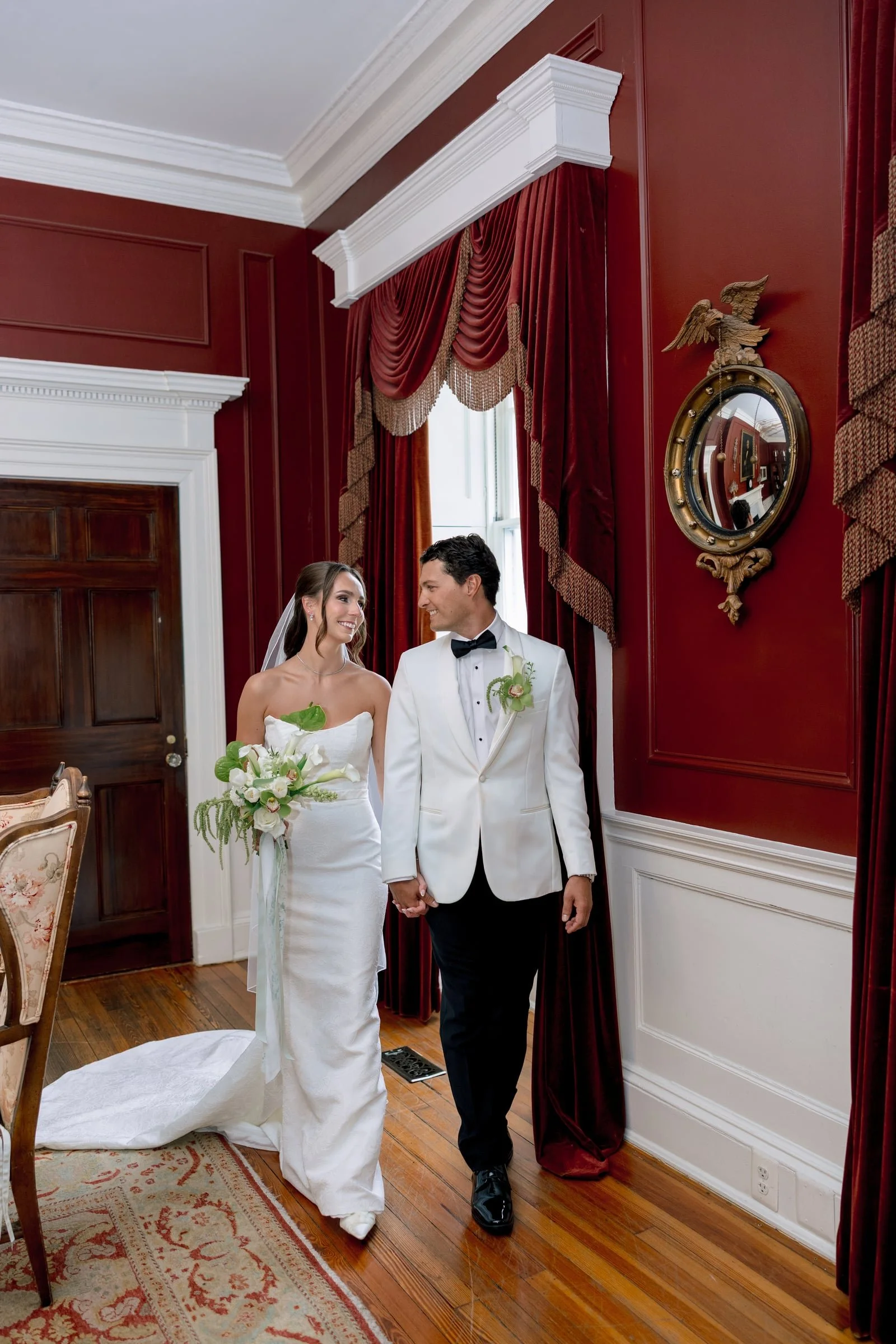 A bride and groom holding hands in a richly decorated room with red walls, white crown molding, heavy red curtains, and a round mirror with an eagle sculpture above it. The bride is wearing a strapless white wedding dress, holding a bouquet, and the groom is in a white tuxedo with a black bow tie.