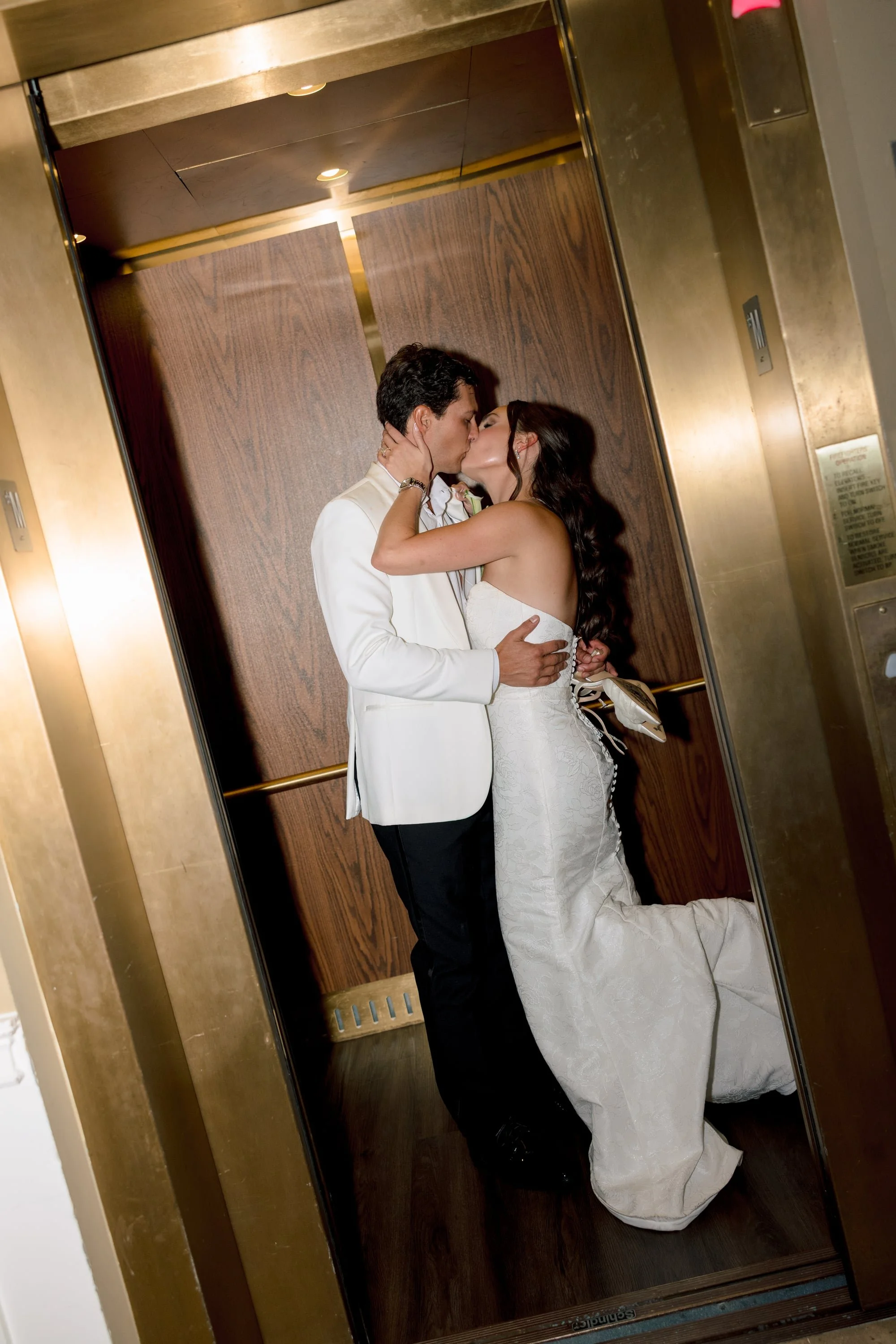 A couple is kissing in an elevator. The man is wearing a white suit and the woman is wearing a white wedding dress. The elevator has wooden walls and is reflected in the mirror.