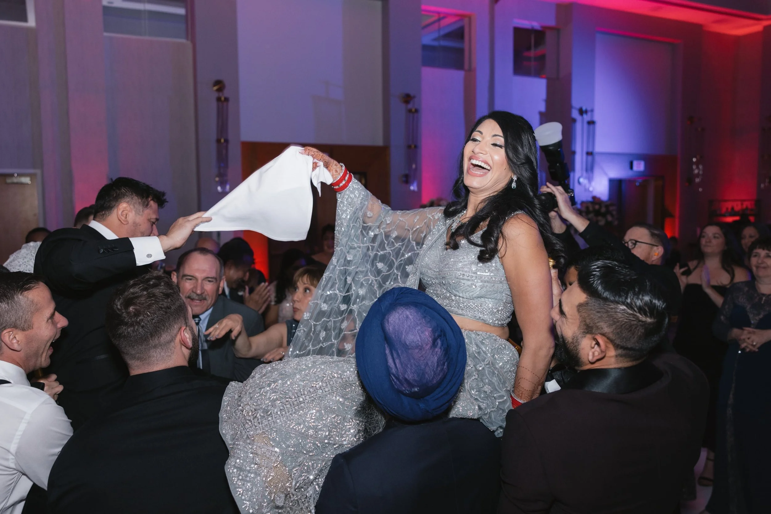 A woman in a silver dress is sitting on a man's shoulders during a festive celebration, surrounded by smiling guests in an elegant venue with purple and red lighting.