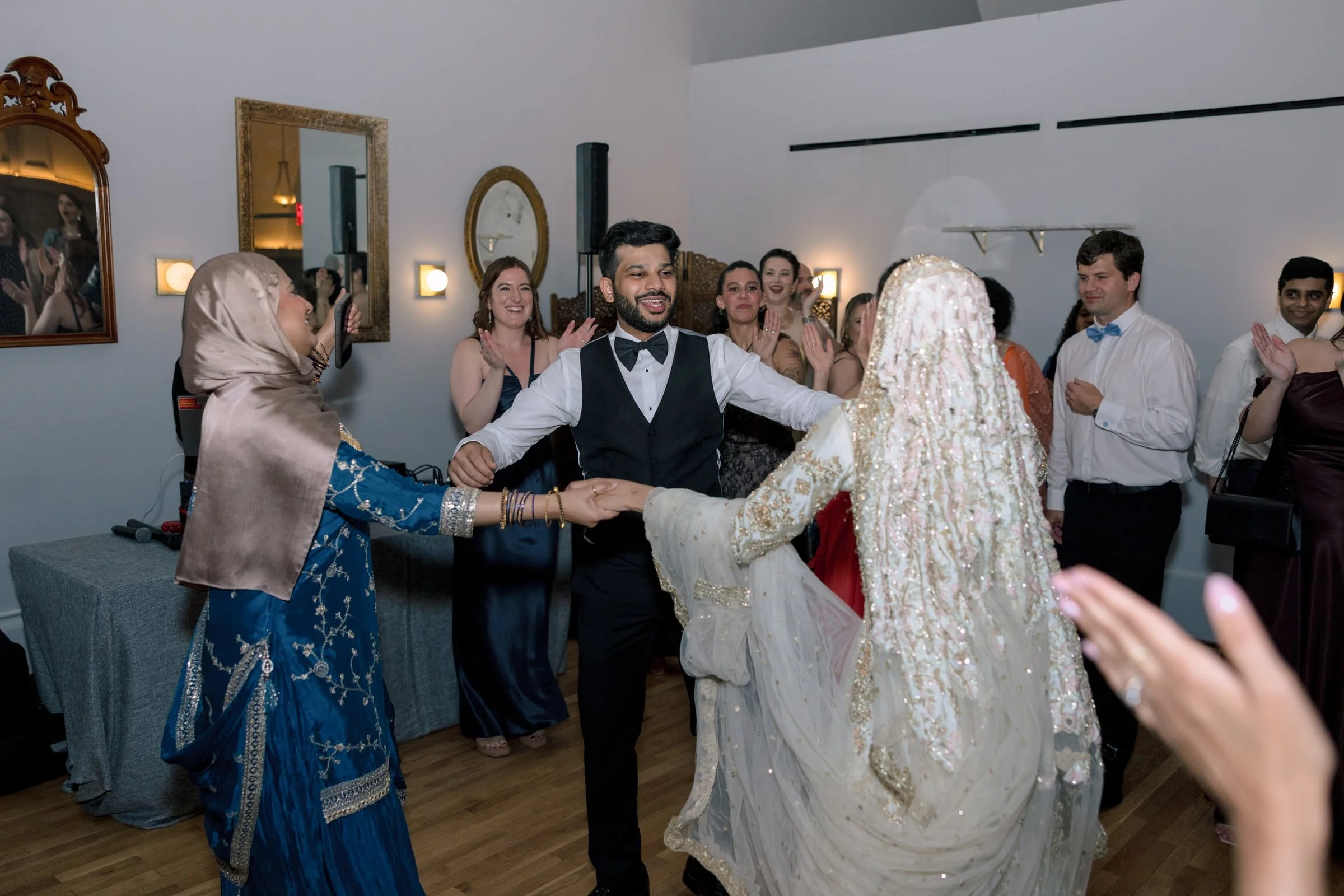 A joyful wedding celebration with a bride in a traditional dress and groom in a tuxedo holding hands, surrounded by smiling guests clapping and enjoying the moment in a decorated indoor venue.