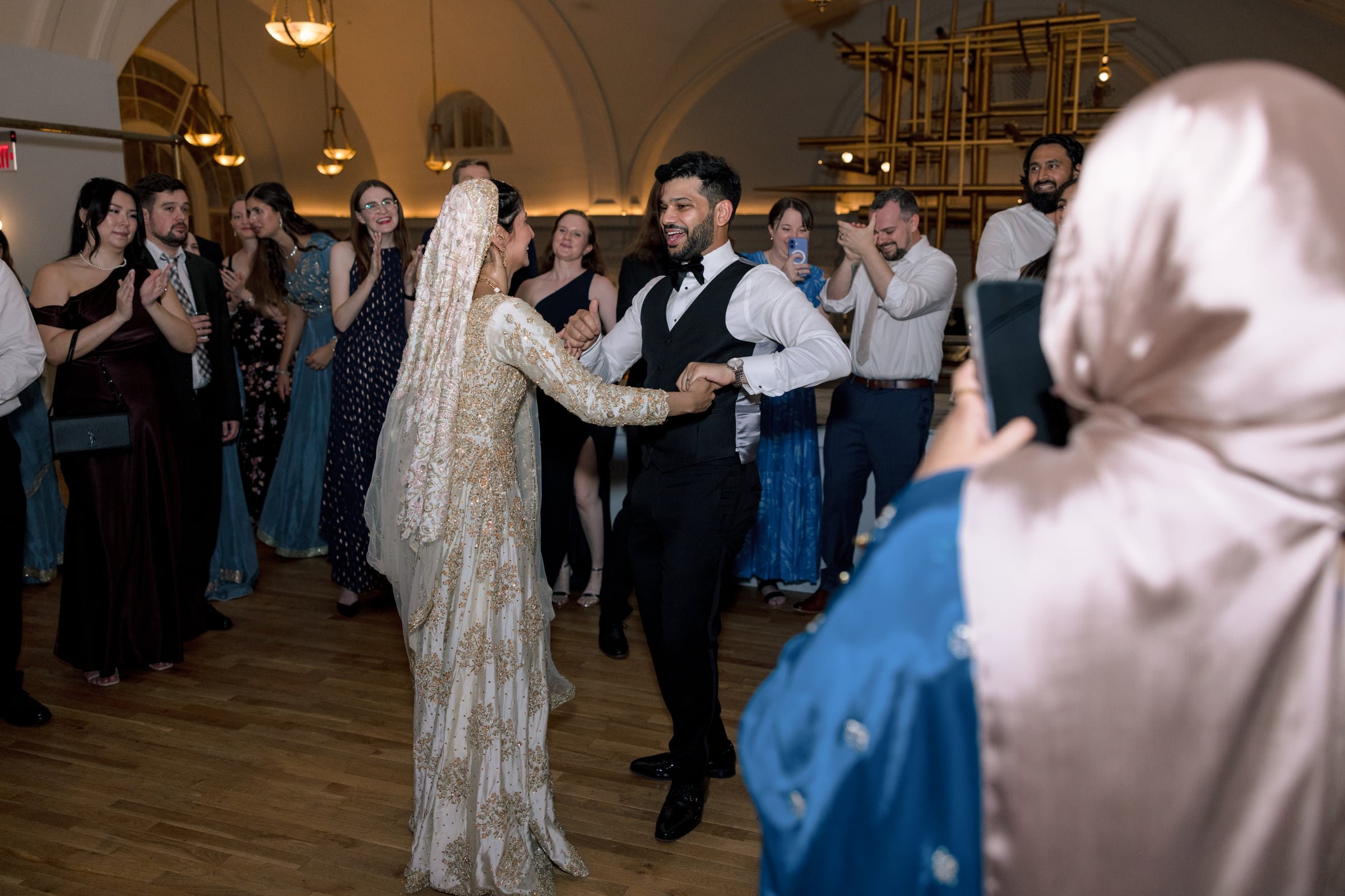 A bride and groom are dancing together at their wedding reception, surrounded by guests, in an elegant indoor venue with high ceilings and warm lighting.
