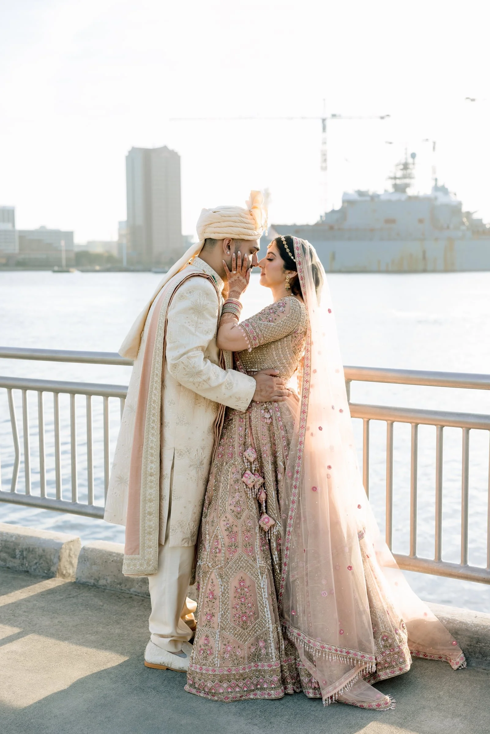 A couple dressed in traditional Indian wedding attire standing close together near a waterfront with a city skyline and a large ship in the background.