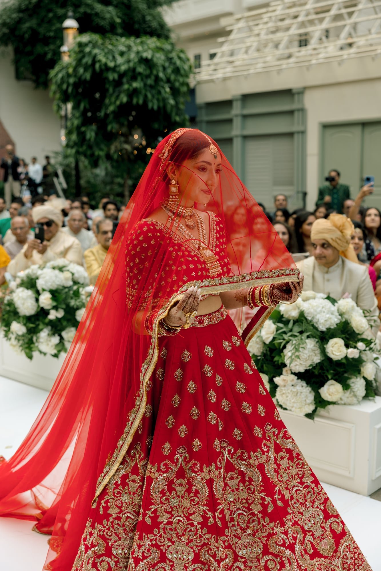 An Indian bride in a red bridal attire with gold embroidery, jewelry, and a red veil, holding a tray in front of a crowd and floral decorations.
