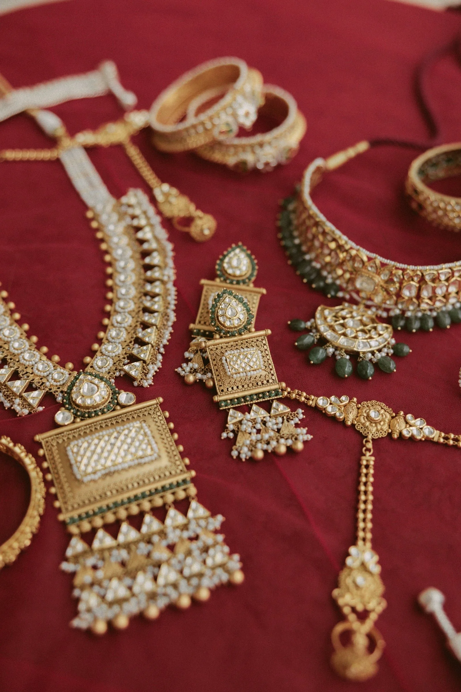 Gold jewelry including necklaces, earrings, and bangles with intricate designs and gemstone embellishments displayed on a red fabric background.