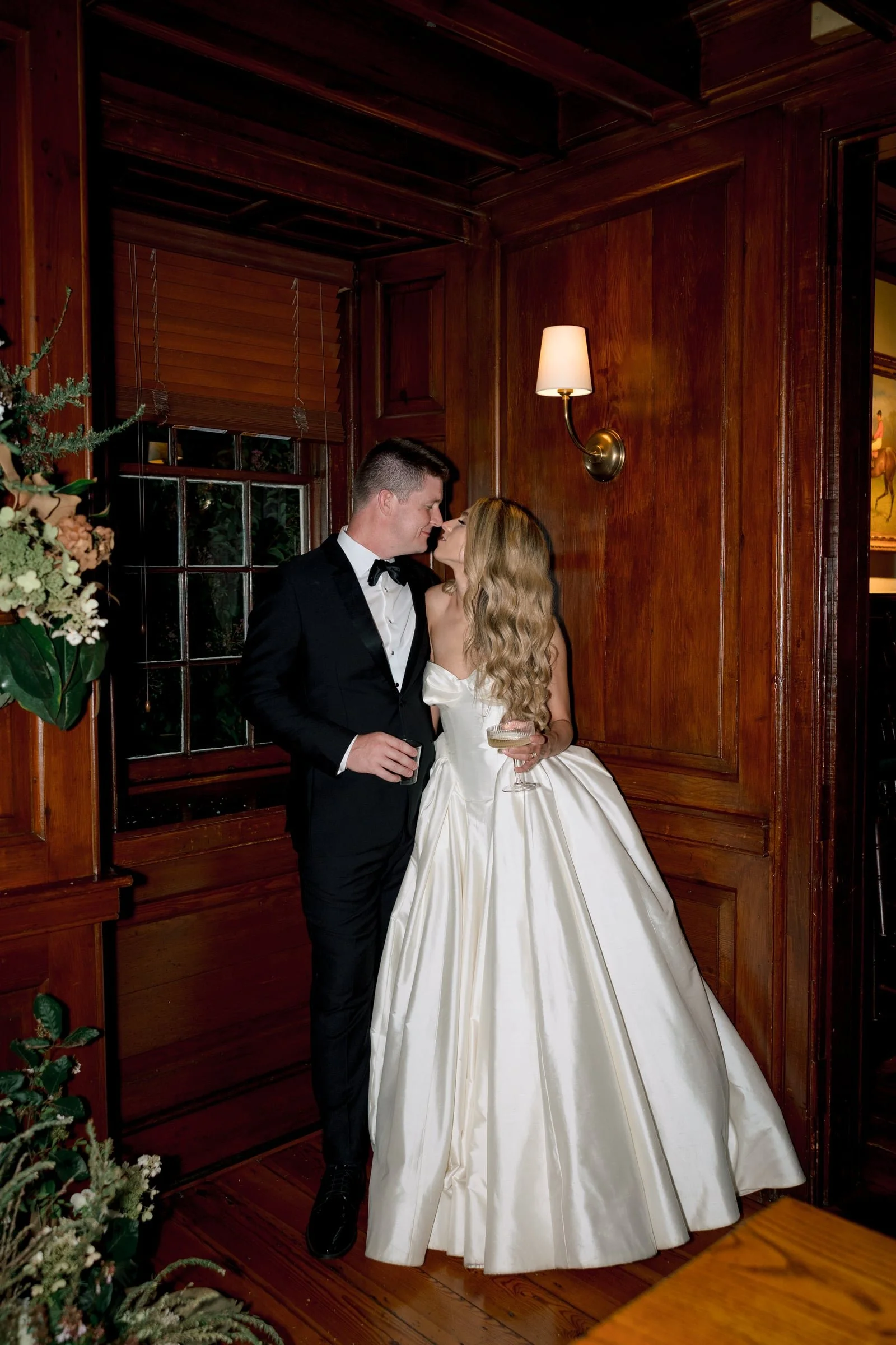 A newlywed couple sharing a romantic moment indoors, with wood-paneled walls, a wall-mounted lamp, a window with blinds, and a floral arrangement nearby.
