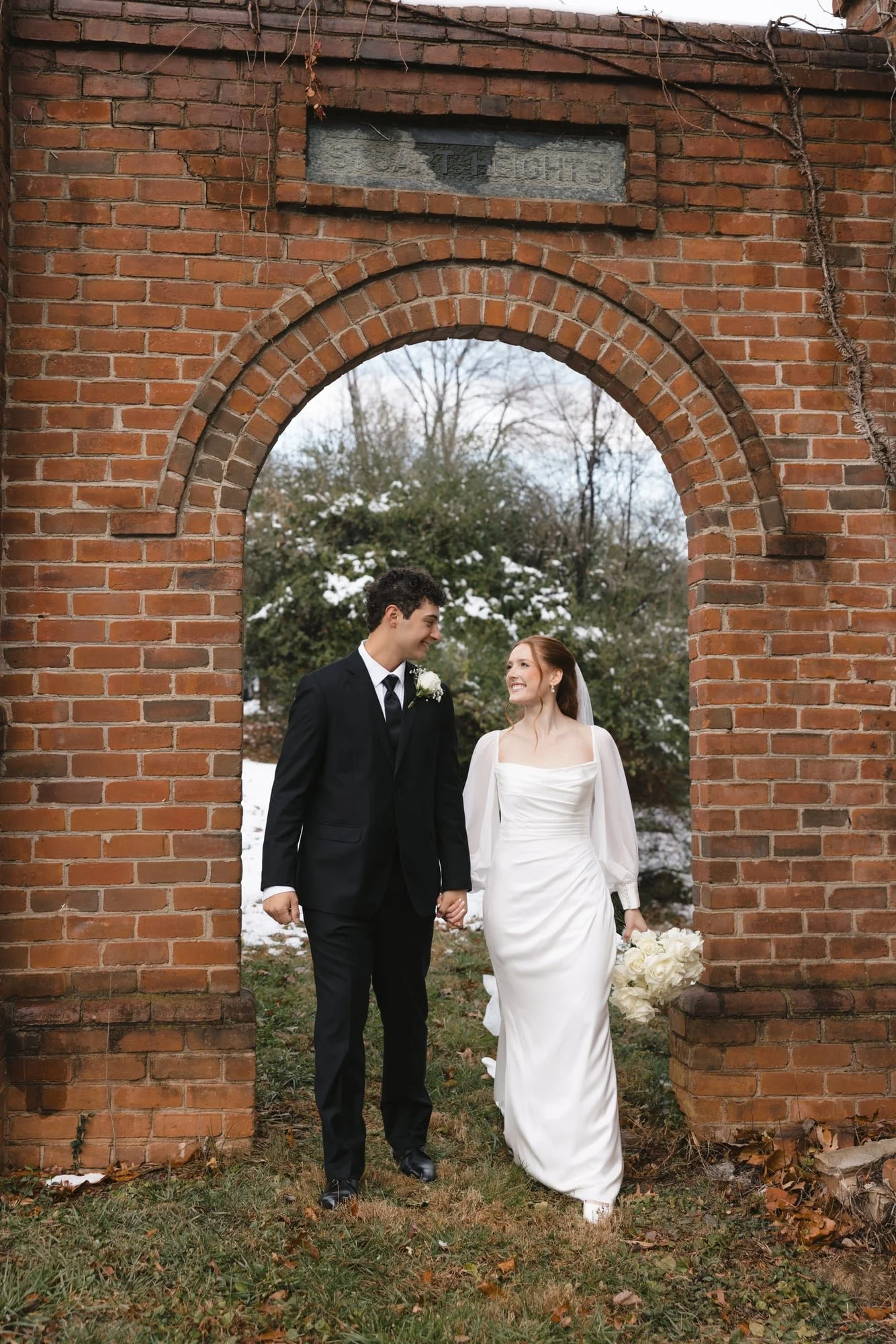 A bride and groom holding hands and walking outdoors through an arched brick structure on a winter day. The bride wears a white wedding dress and carries a bouquet, and the groom wears a black suit and tie. They are smiling at each other, in front of