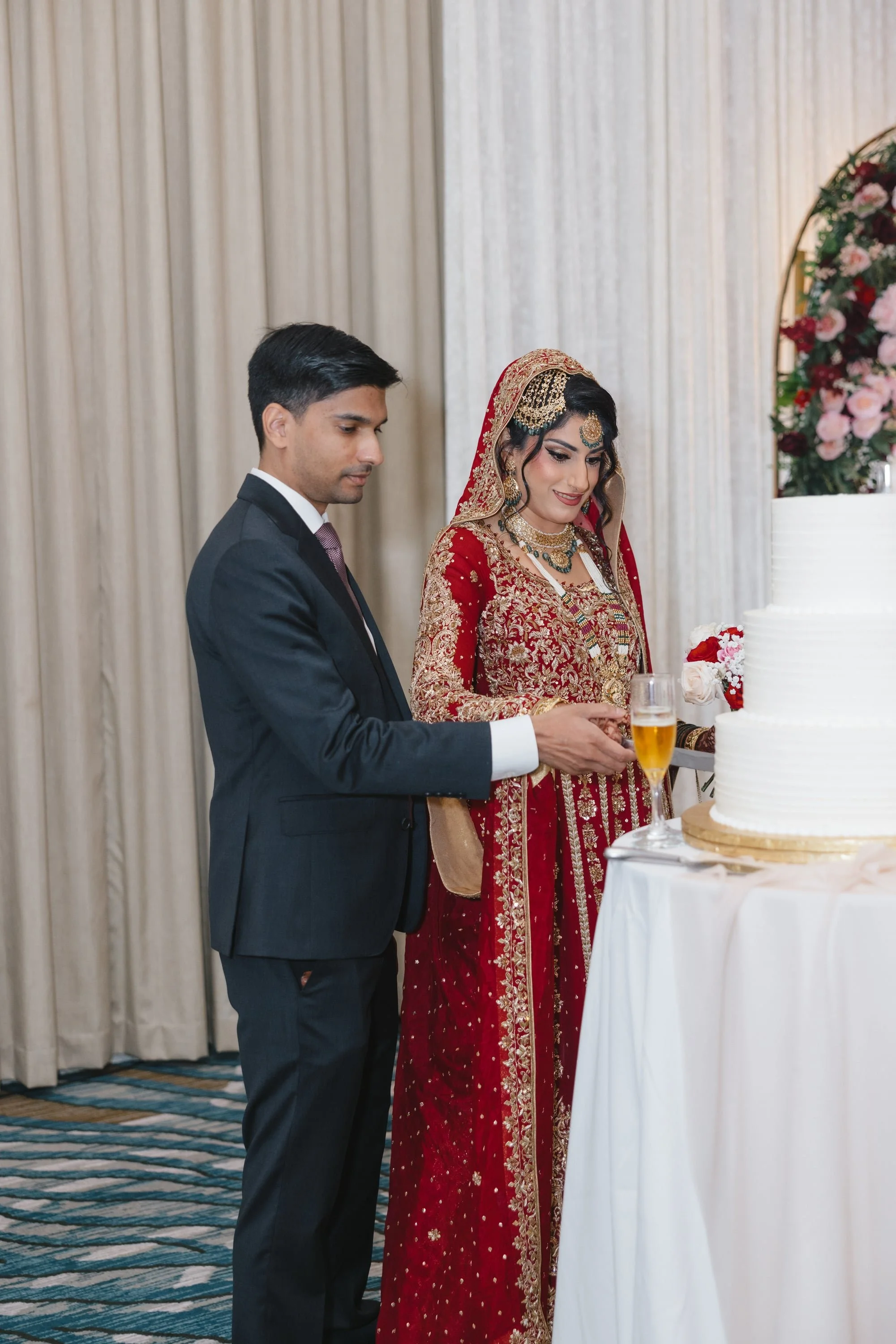 A groom in a dark suit and a bride in a red and gold traditional South Asian wedding dress cutting a white wedding cake with floral decorations, standing indoors with cream-colored curtains in the background.