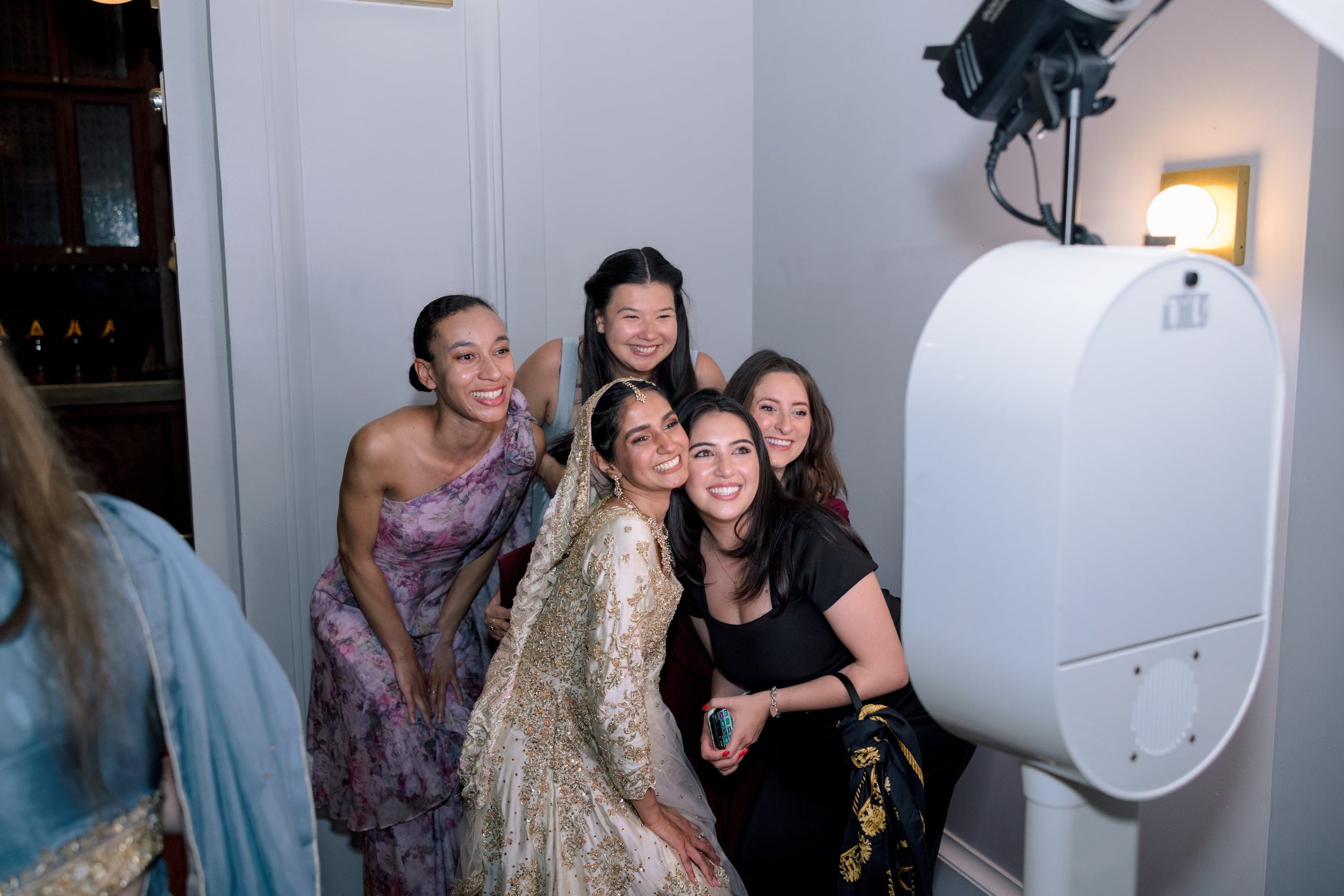 A group of five women taking a selfie in front of a large medical imaging machine, possibly an MRI or CT scanner, in a clinical setting. They are smiling and dressed in formal or traditional attire.