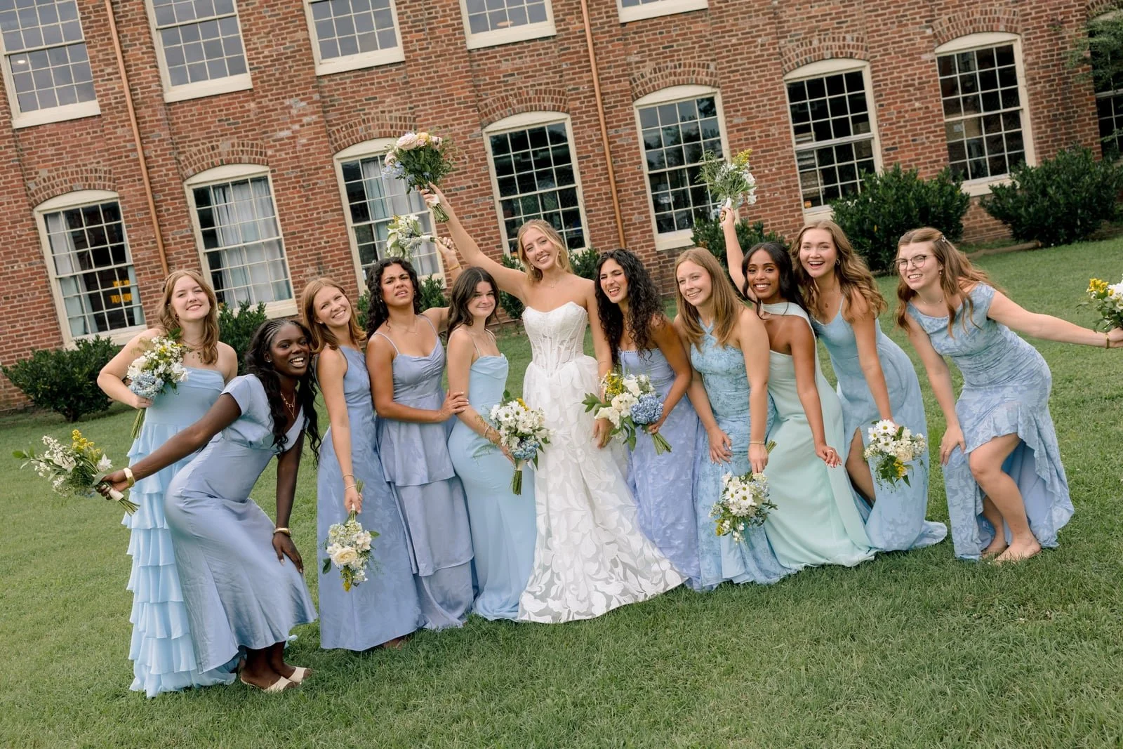 A bride in a white wedding dress surrounded by ten bridesmaids in pastel blue dresses, outdoor wedding setting in front of a brick building, all holding bouquets, celebrating and smiling.