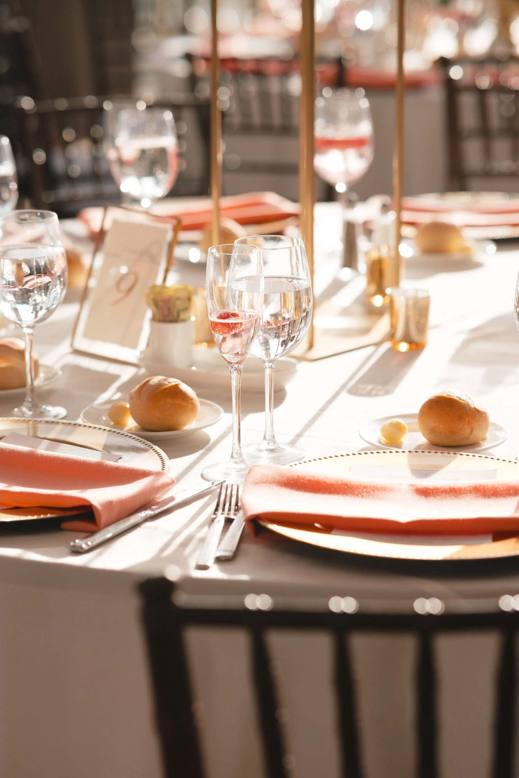 A well-set dining table with pink napkins, glassware, bread rolls, and place settings, illuminated in warm sunlight, in a dining area with blurred background.
