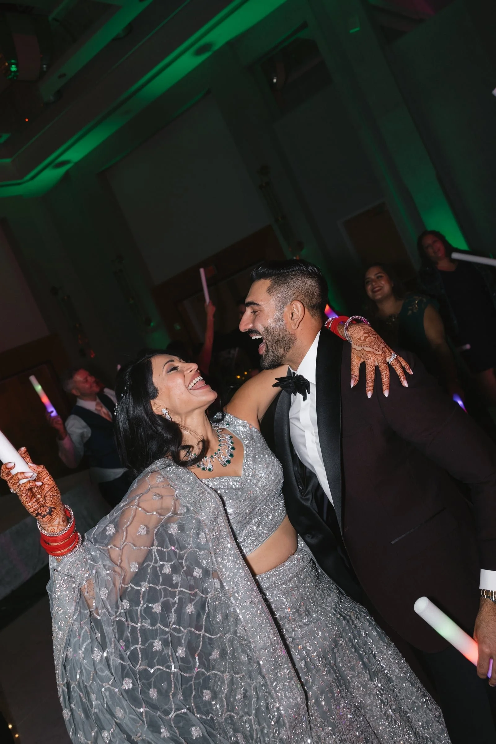 A joyful couple dancing at a celebration, surrounded by guests, with colorful lighting and decorations.