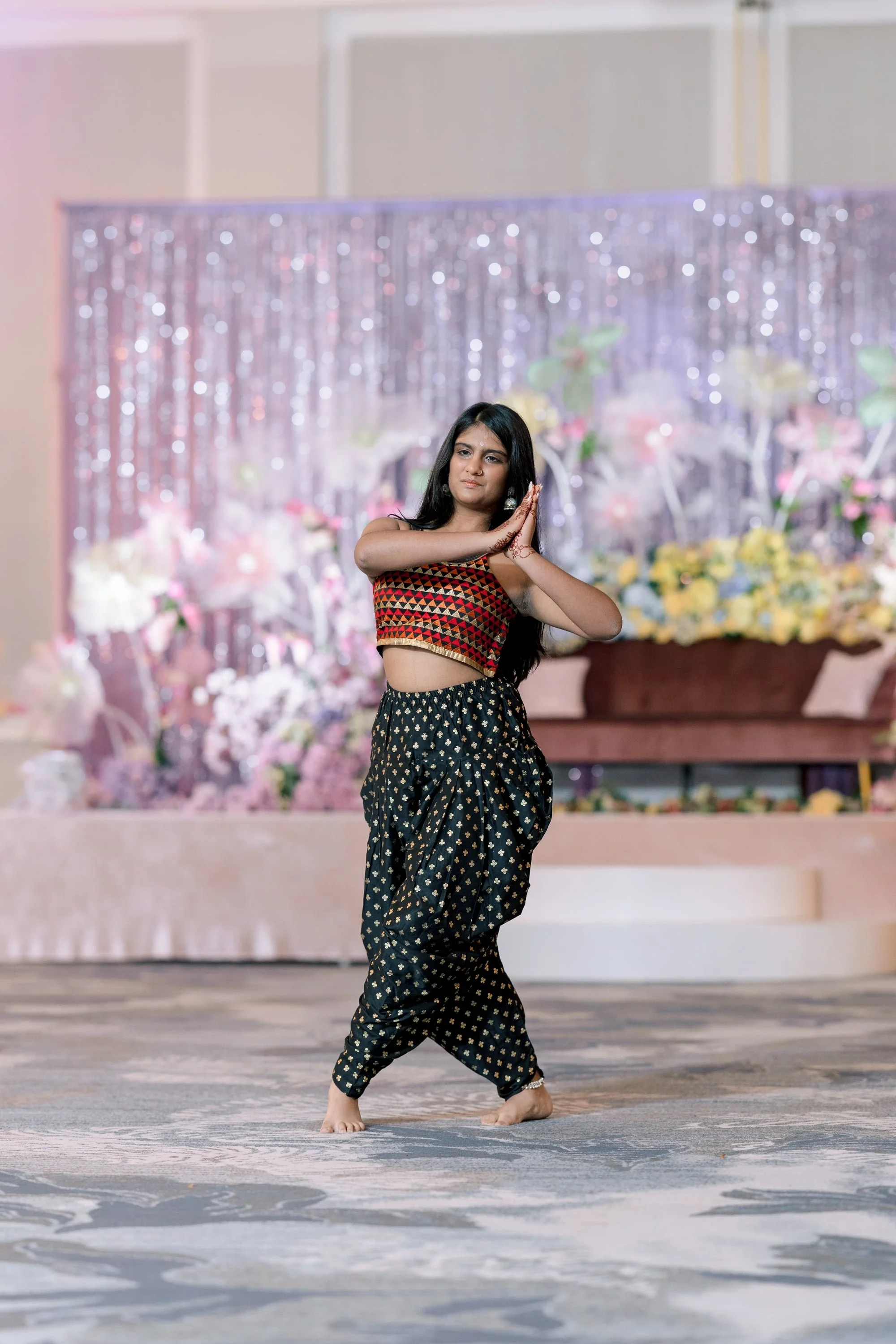 A young woman in traditional Indian attire performing a dance, standing barefoot on a decorated stage with floral arrangements and a sparkling backdrop.