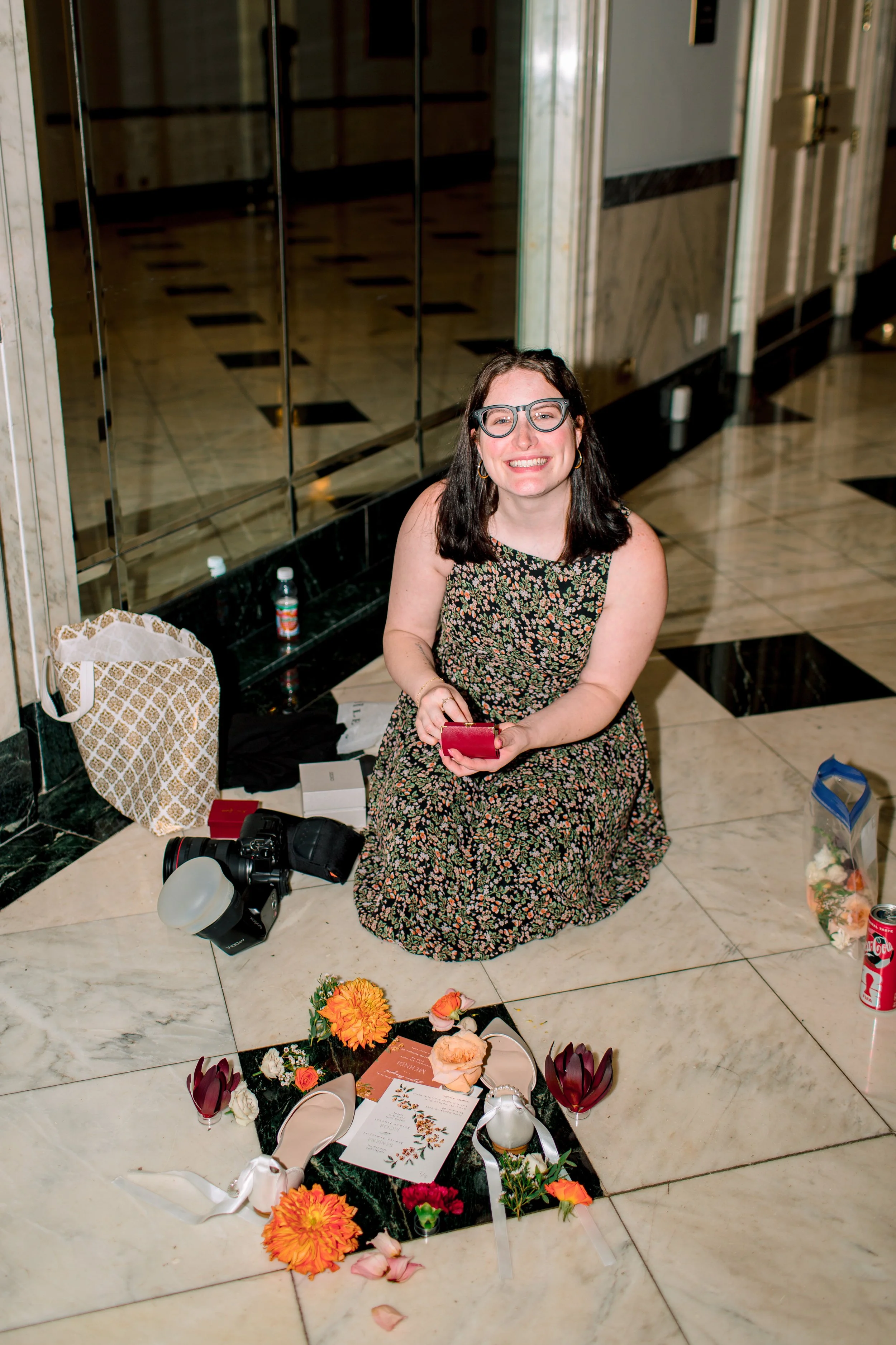 A woman with dark hair, glasses, and earrings is sitting on a marble floor in front of a memorial display with flowers, candles, and programs. She is smiling and holding a small red box. The background is a hallway with marble walls and reflective surfaces.