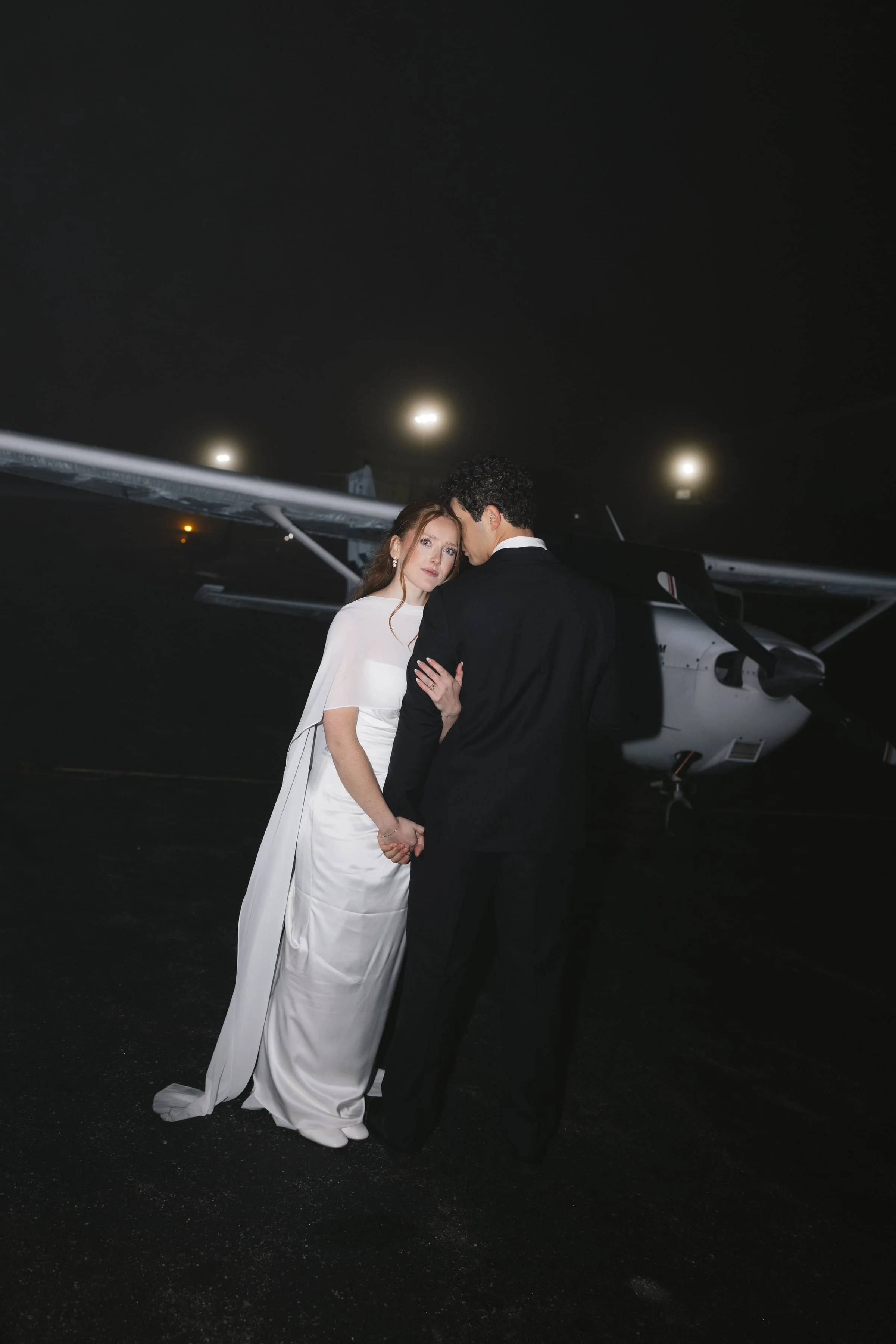 A couple dressed in wedding attire holding hands at night near a small private jet, with the woman in a white wedding gown and the man in a black tuxedo.