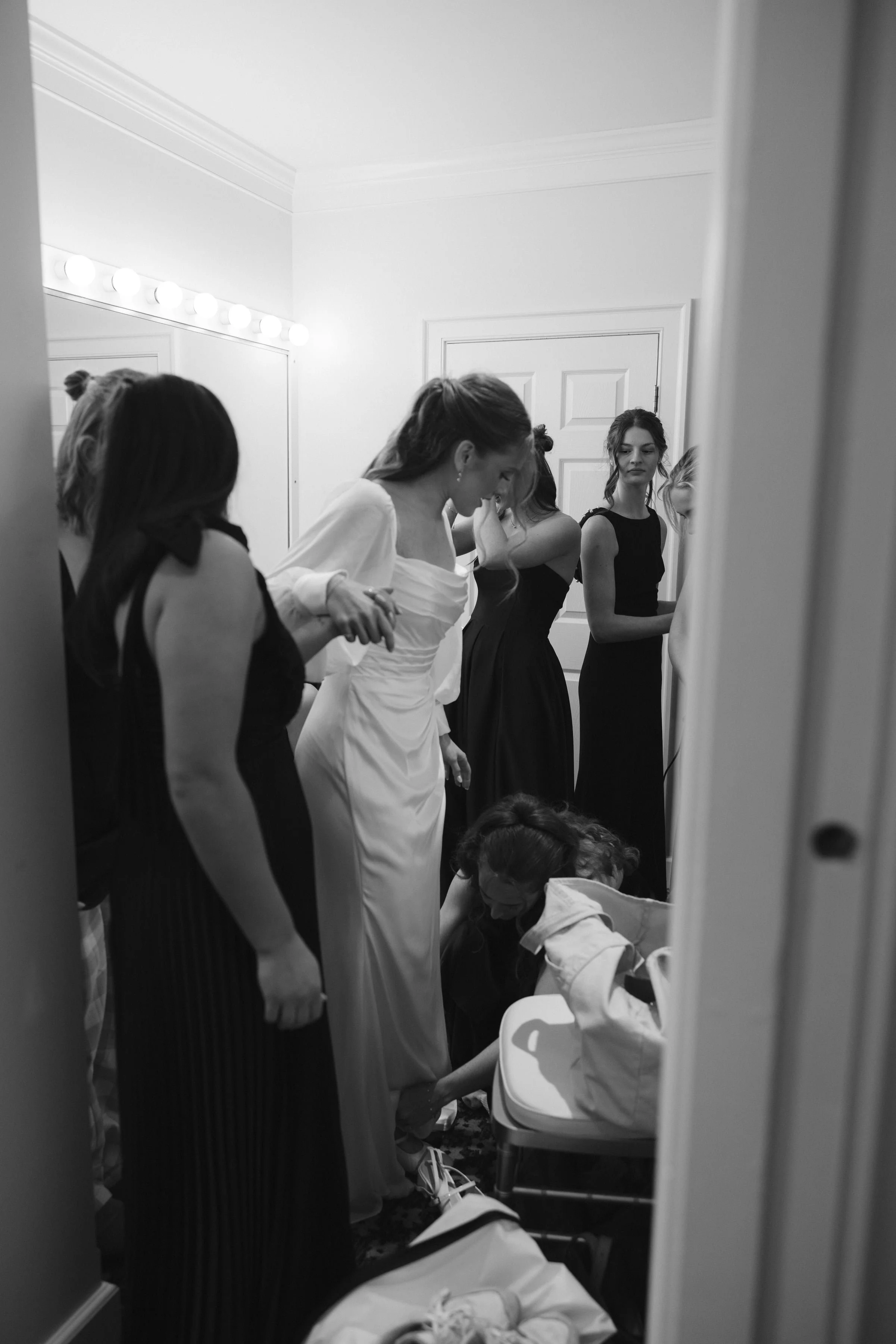Women preparing for an event in a dressing room, with one woman in a white dress and others in black dresses, and a changing station or makeup mirror visible.