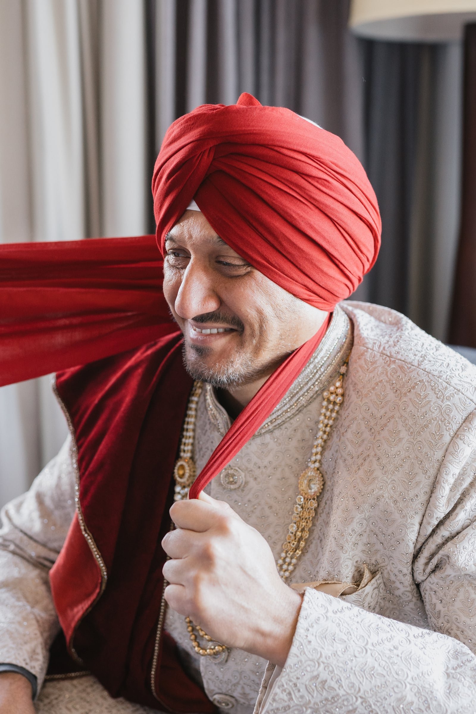 A man wearing a red turban and traditional cream-colored clothing adorned with gold jewelry, smiling and holding the scarf of his turban.