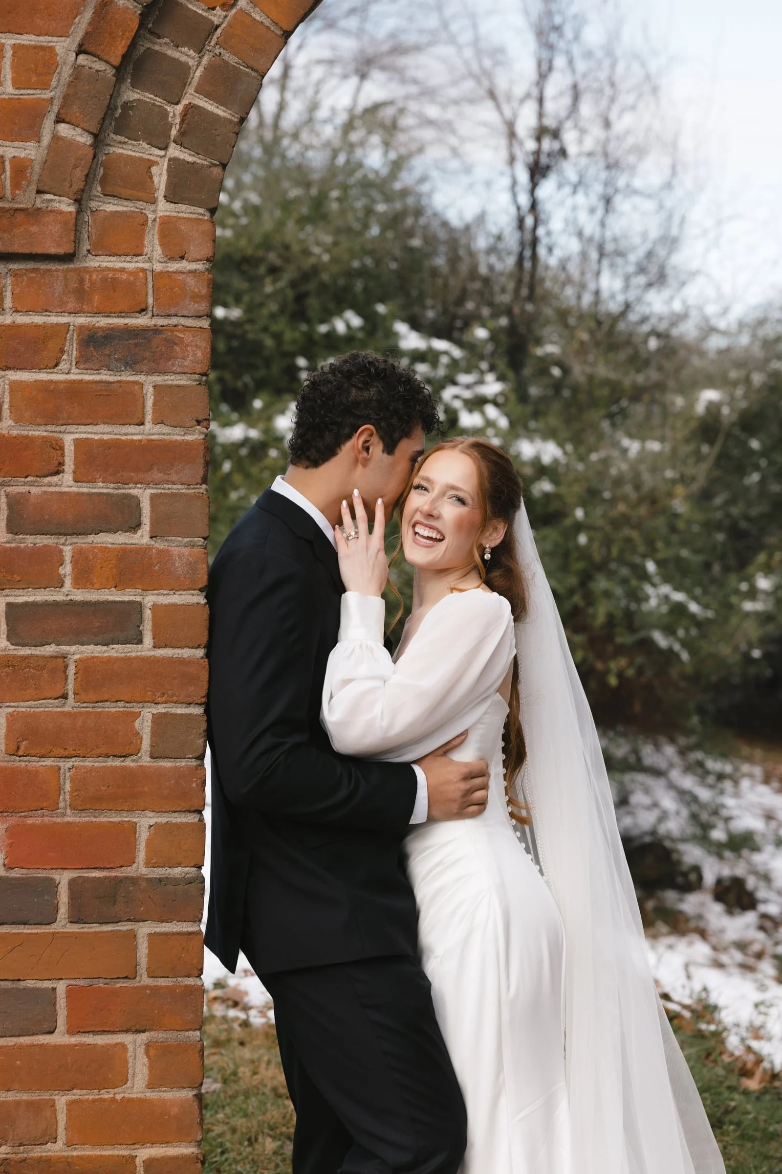 A bride and groom laughing and embracing outdoors near a brick wall, with snowy trees in the background.