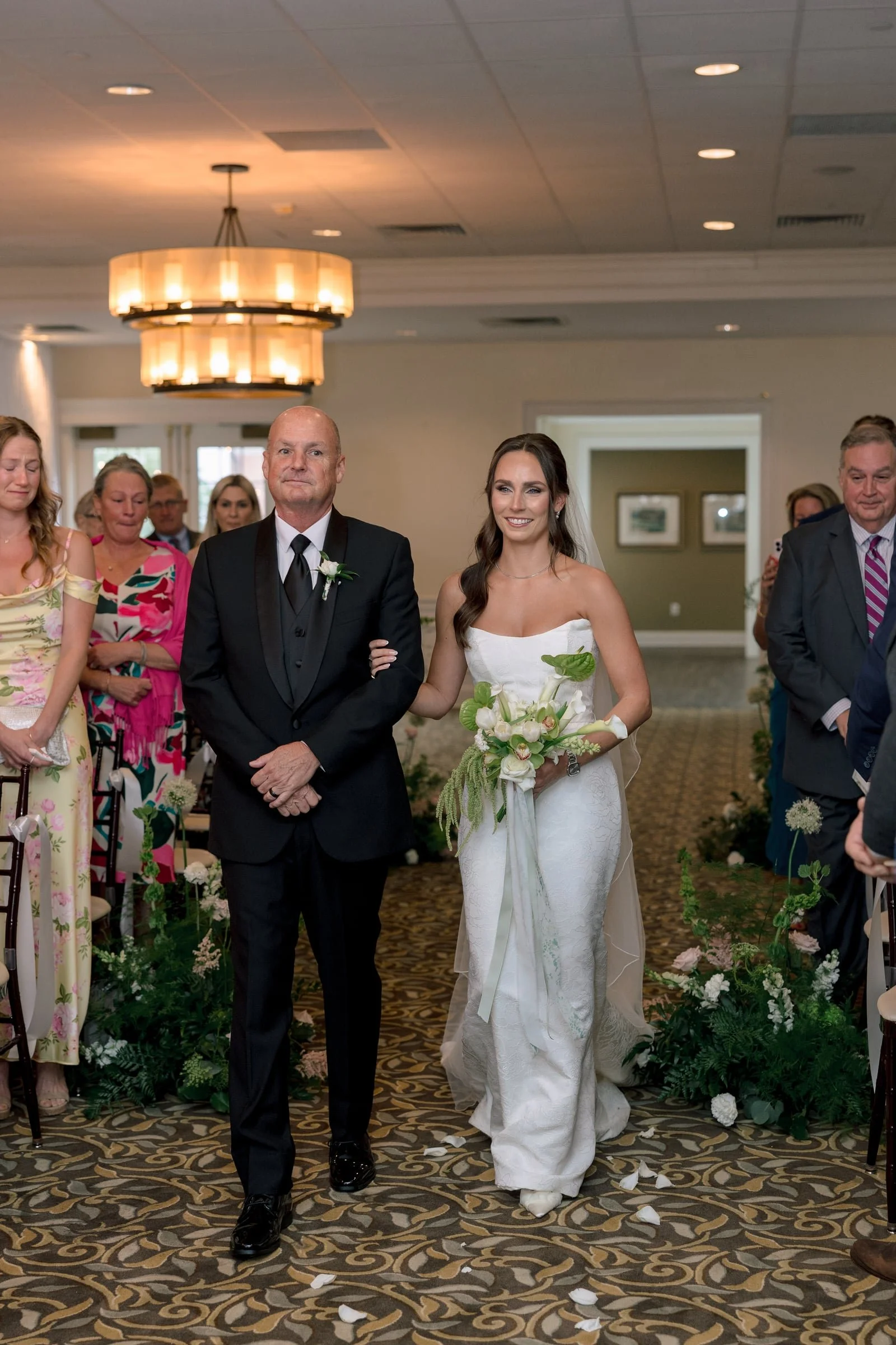 A bride and her father walking down the aisle during their wedding ceremony in a decorated indoor venue, surrounded by seated guests and floral arrangements.