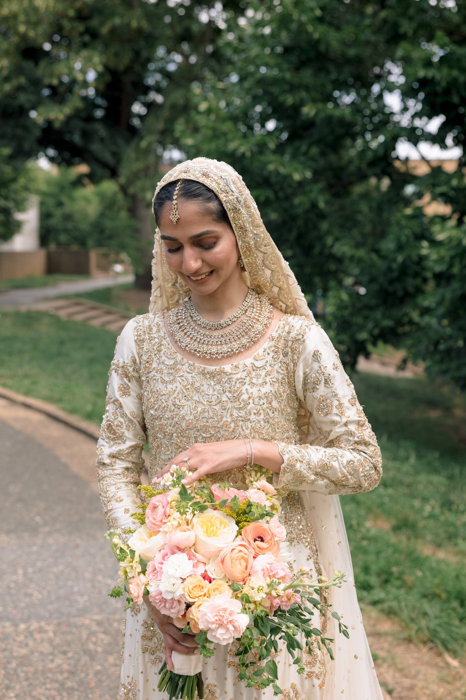 A woman in a traditional, ornate wedding dress holding a large bouquet of pink and cream flowers, standing outdoors with trees in the background.