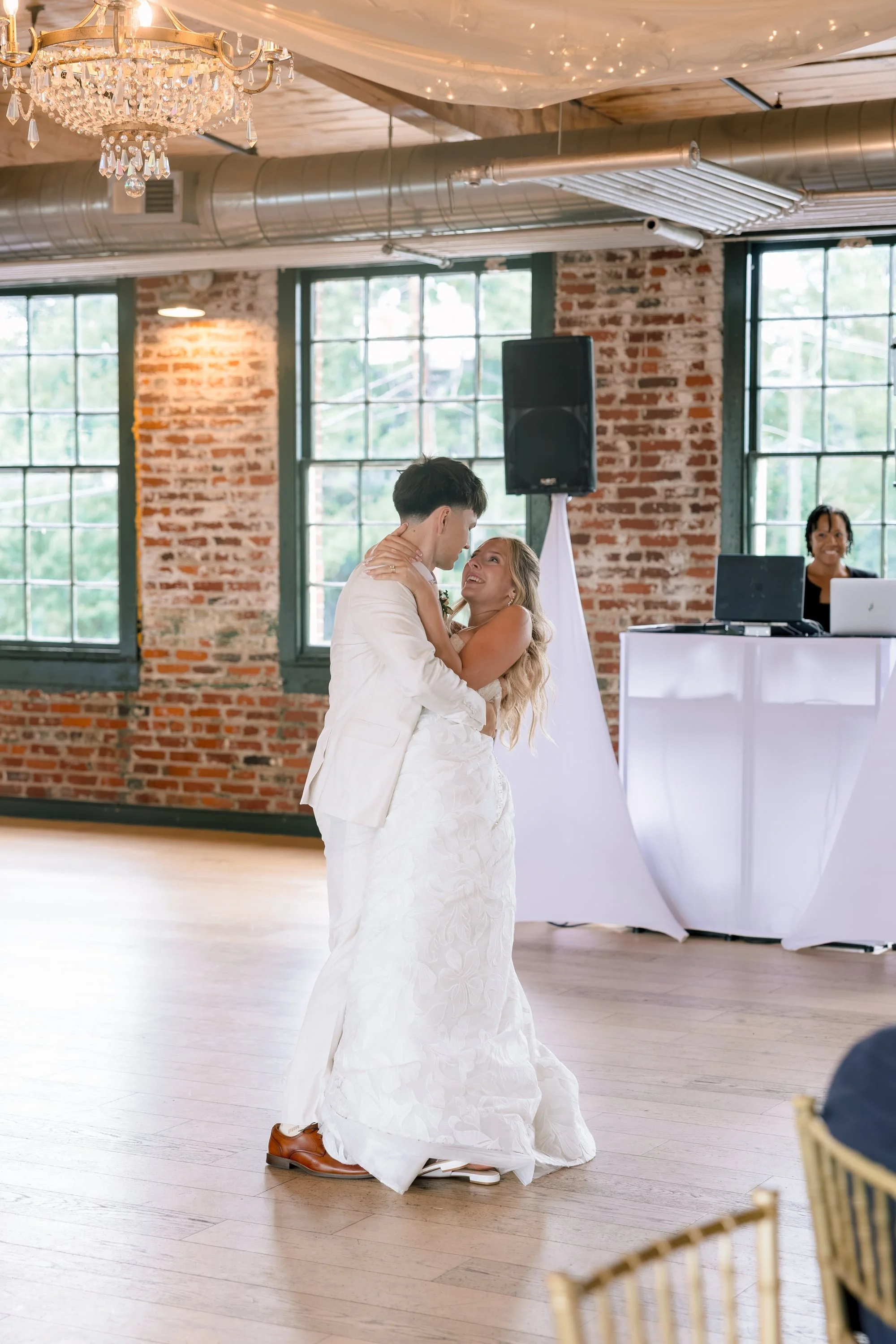 A bride and groom sharing a dance at their wedding reception in a venue with brick walls and large industrial-style windows. The bride wears a white wedding gown and the groom a white suit. A woman stands behind a DJ booth, and there are empty chairs