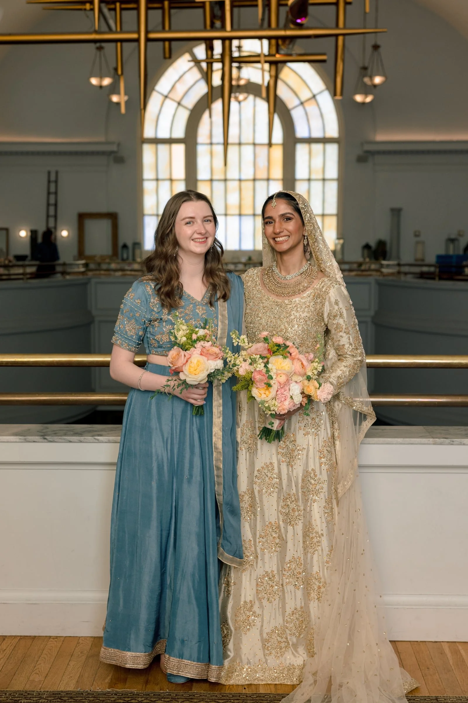 Two women, one in a blue traditional dress and the other in a cream and gold embellished traditional dress, smiling and holding bouquets of flowers, standing inside a spacious hall with large arched window and a decorative gold chandelier overhead.