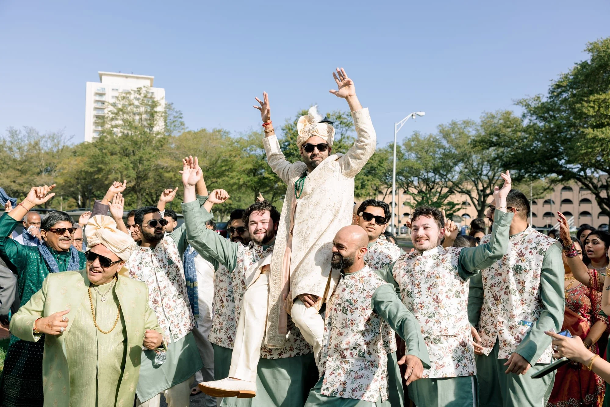 A group of people celebrating outdoors during daytime, with a man dressed in traditional Indian attire riding on the shoulders of others. The crowd appears joyful with many raising their hands, set against a backdrop of trees and a tall building unde