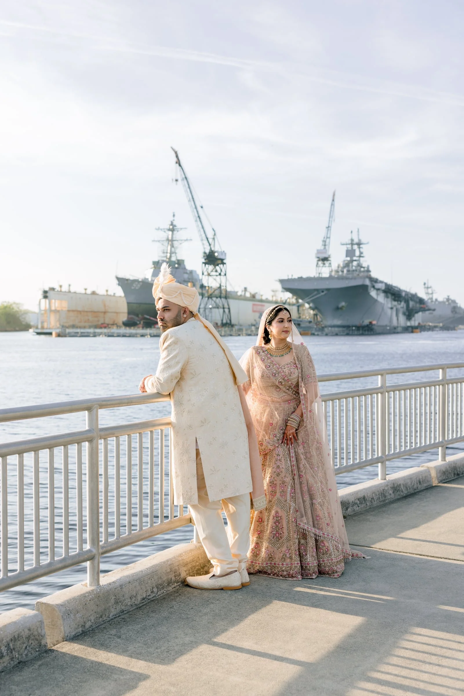 A newlywed Indian couple in traditional wedding attire standing on a waterfront promenade with ships and cranes in the background.