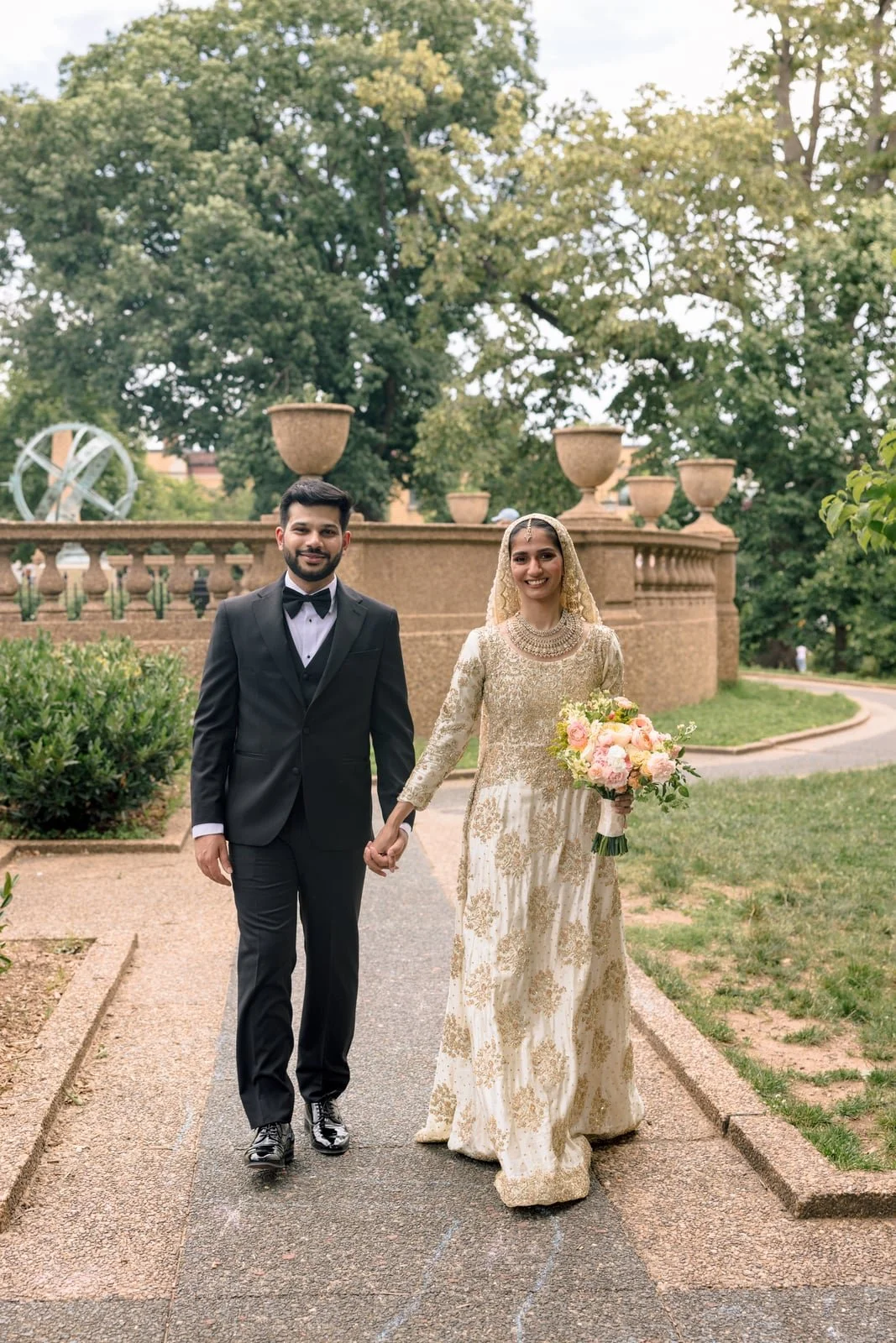 A wedding couple walking hand-in-hand outdoors, with a man in a black tuxedo and a woman in an ornate traditional bridal dress holding a bouquet of pink and white flowers, behind them a stone wall and green trees.