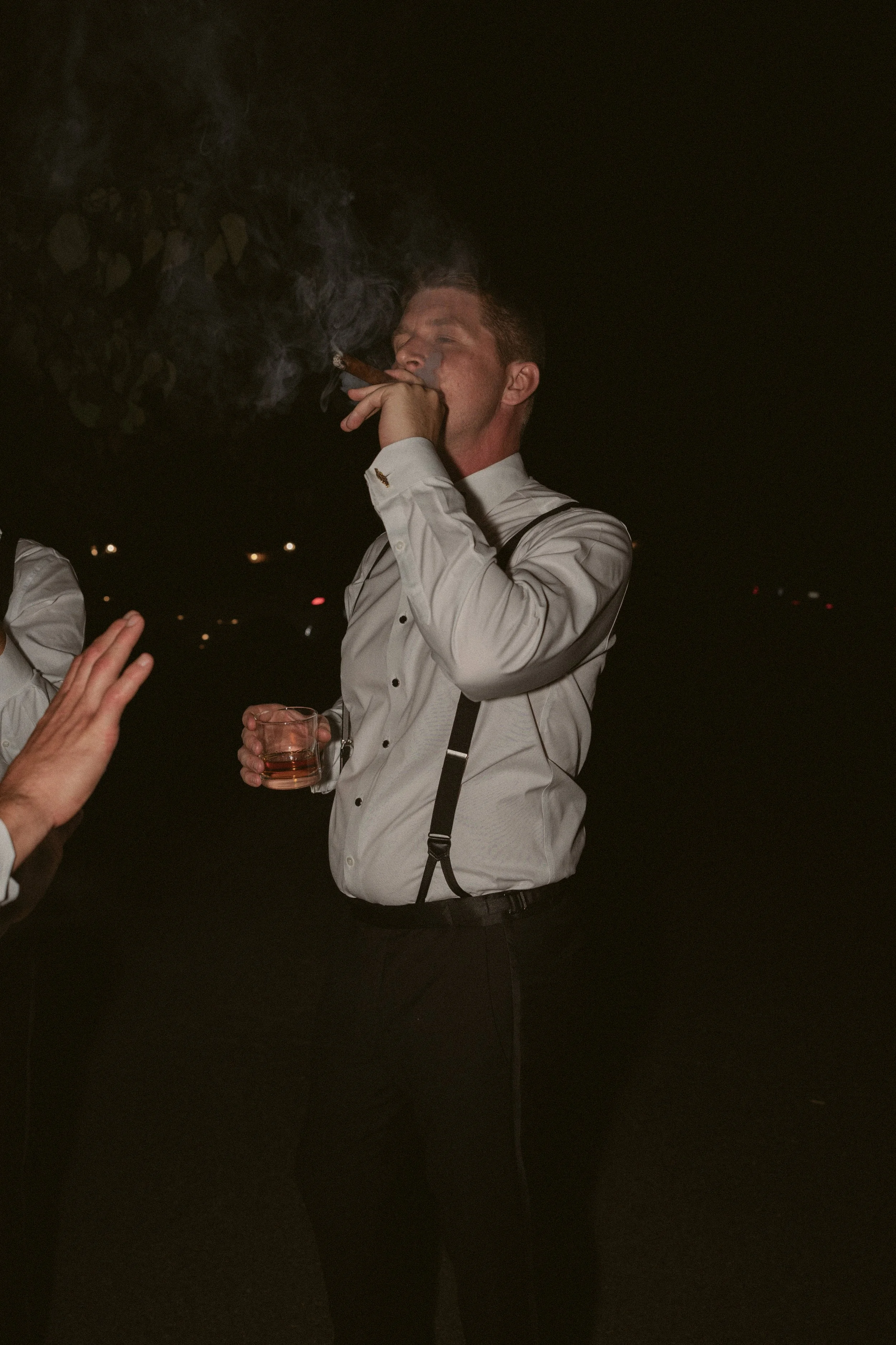 A man in formal attire, holding a drink in one hand and a cigar in the other, is smoking outdoors at night.