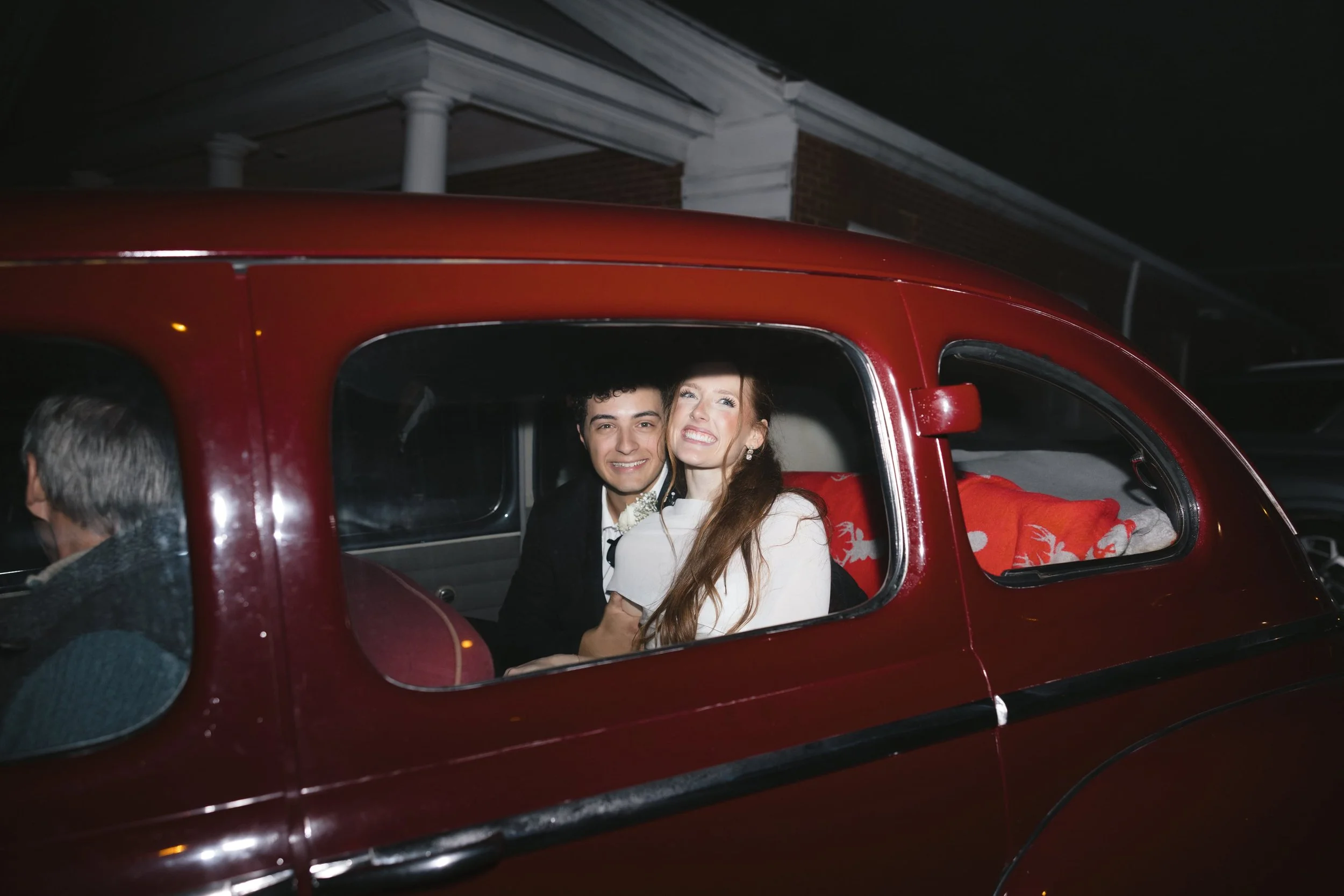 A couple sitting inside a red vintage car at night, smiling and looking out the window. A person with gray hair is partially visible outside the car, and a house with white columns and brick walls is in the background.