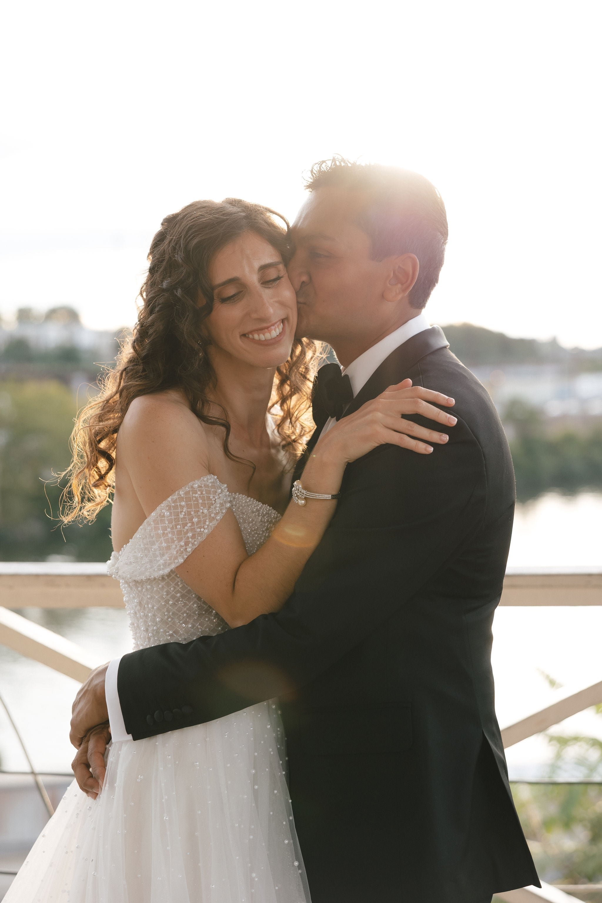 A bride and groom sharing a romantic moment outdoors during sunset, with the groom kissing the bride on the cheek. The bride has curly hair and is smiling with her eyes closed, wearing an off-shoulder wedding dress, while the groom is in a black tuxe