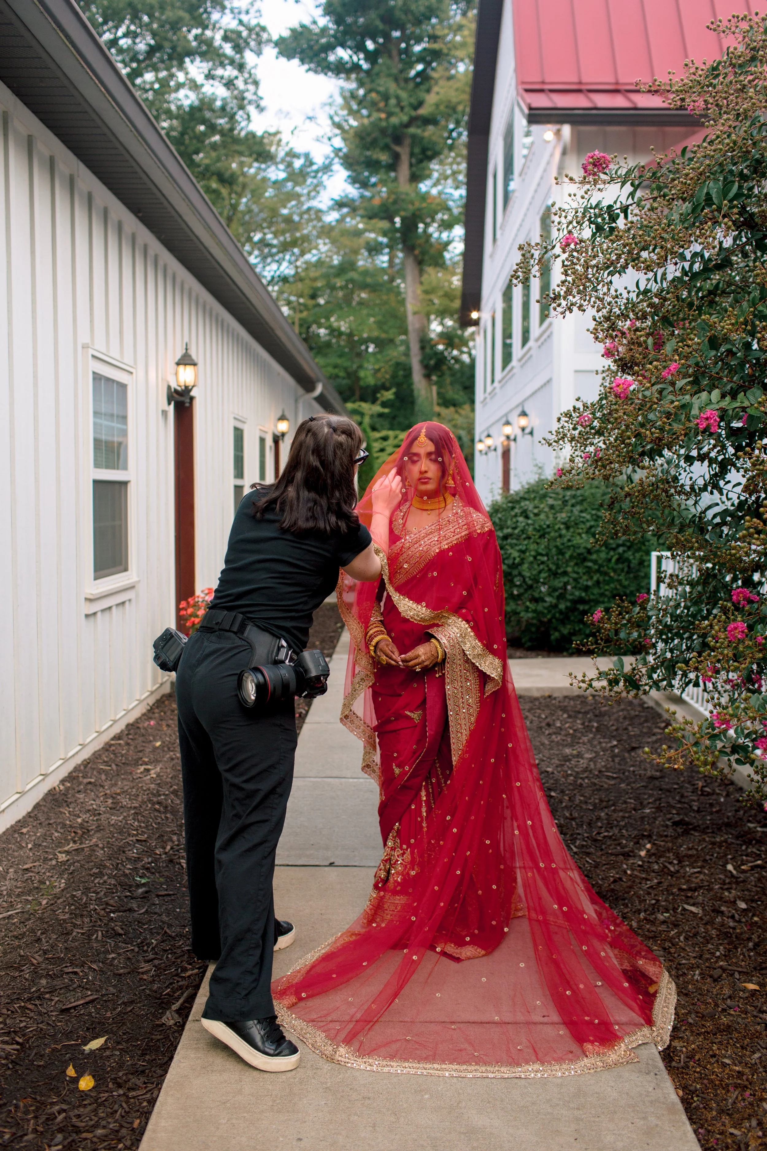 A woman in a red and gold traditional Indian wedding dress and veil is having her makeup done by a makeup artist on a sidewalk outside a white building with pink flowers.