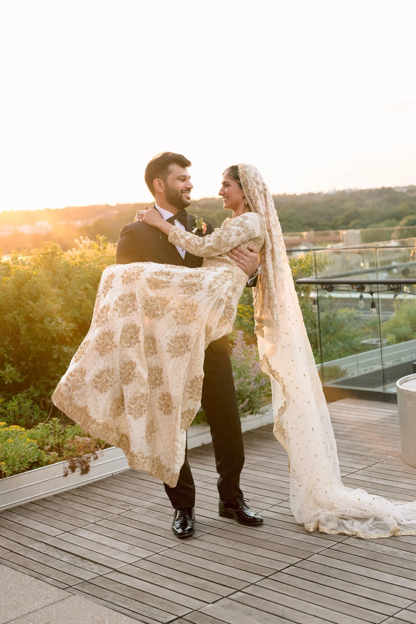 A groom in a black tuxedo holds a smiling bride in a white and gold embroidered wedding dress outdoors on a wooden deck, with a sunset and green landscape in the background.