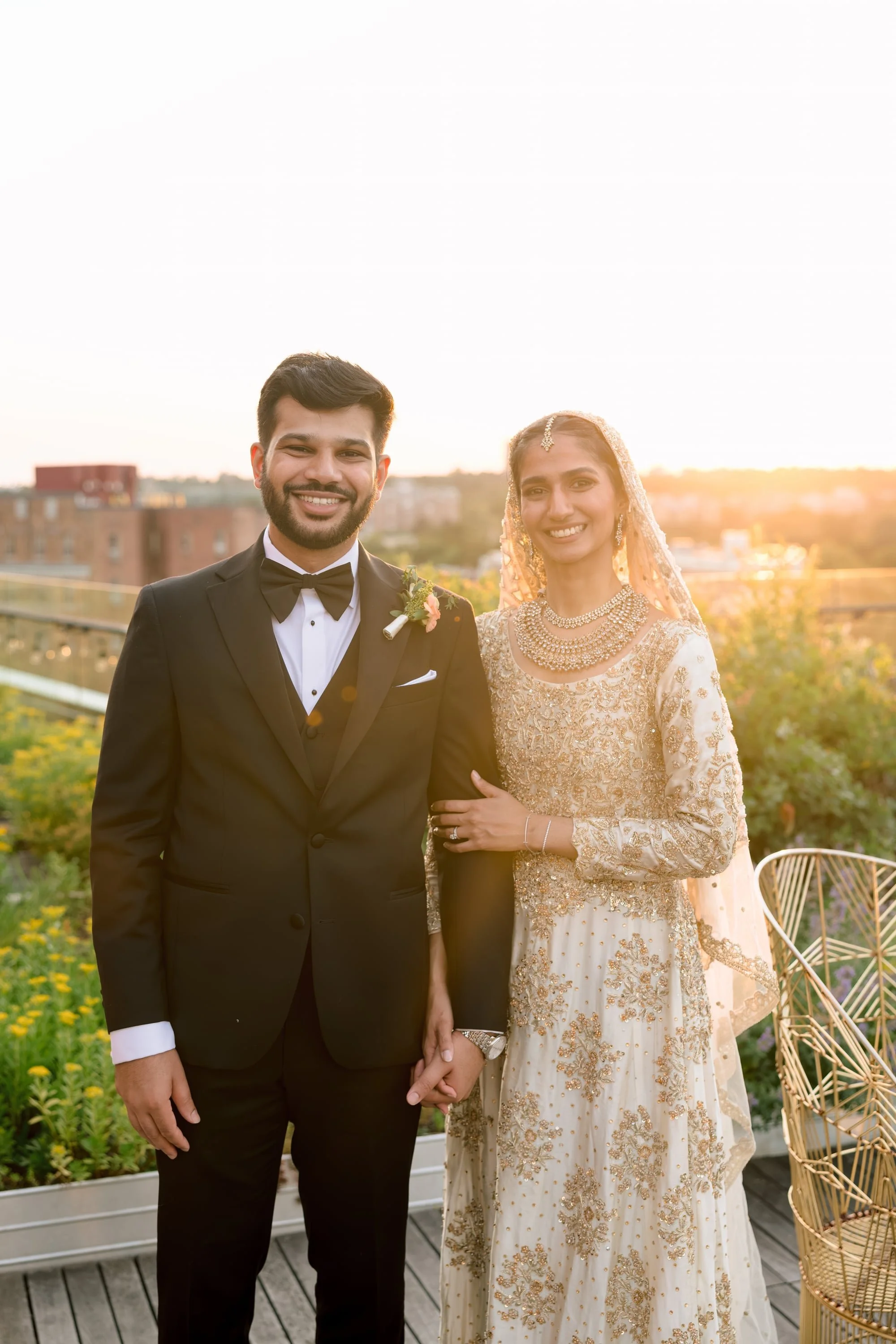 A couple in wedding attire standing outdoors during sunset, holding hands, with city buildings in the background and greenery around.