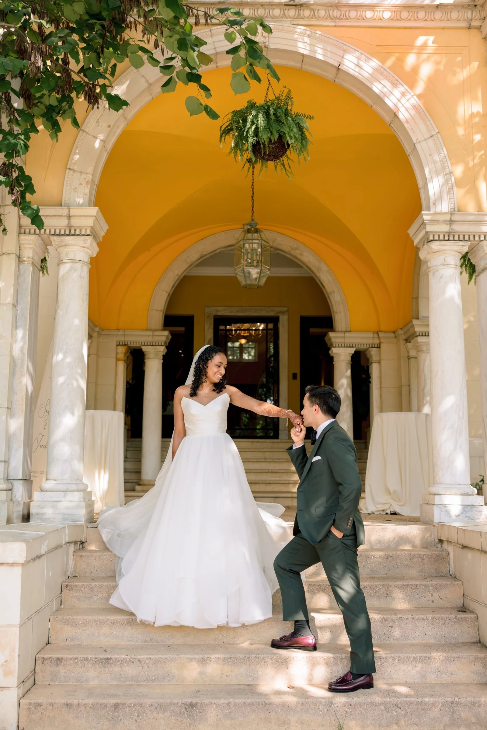 A bride in a white wedding dress and a groom in a dark suit with a green tie are on the steps of an elegant building with yellow walls and white stone columns. The bride is holding the groom's hand as he kneels on one knee. There are green plants and hanging ferns overhead, with a large hanging light fixture and an arched entrance in the background.