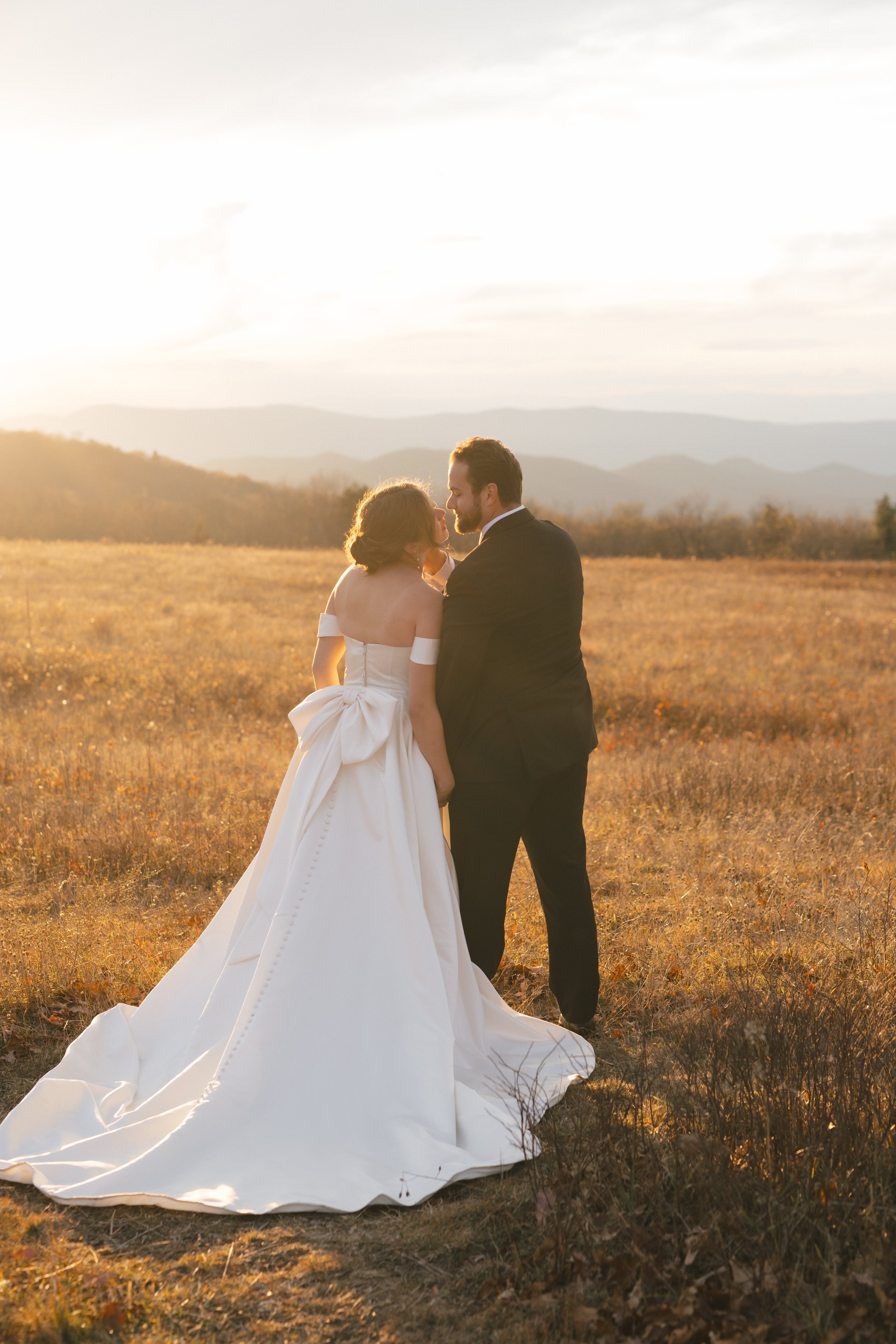 A bride and groom standing close in a field at sunset, with mountains and cloudy sky in the background. The bride wears a white wedding gown with a full skirt, and the groom wears a dark suit. They are gazing at each other affectionately.