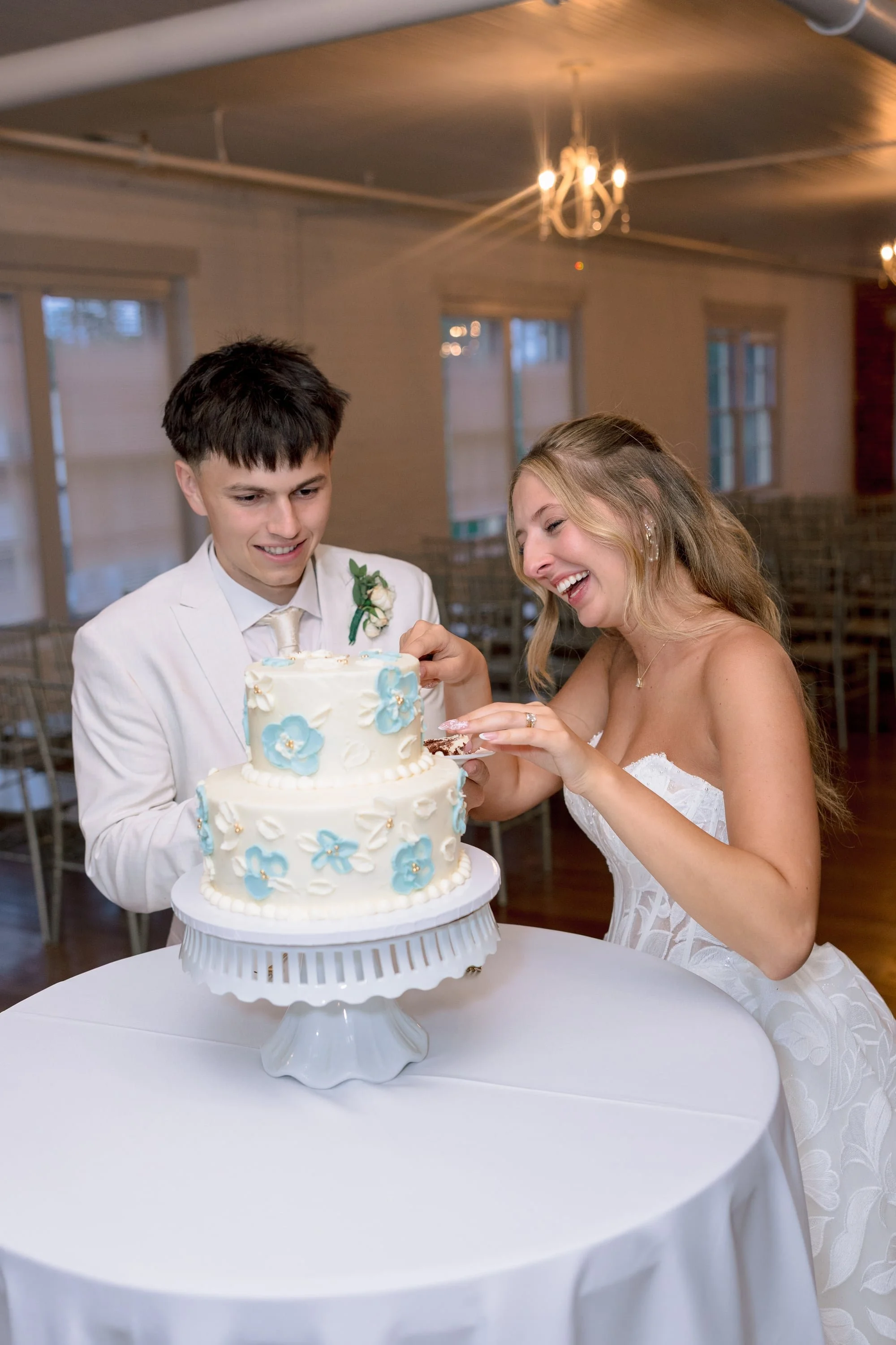 A bride and groom cutting their wedding cake together, standing behind a round table covered with a white cloth, inside a decorated indoor reception hall with hanging light fixtures.