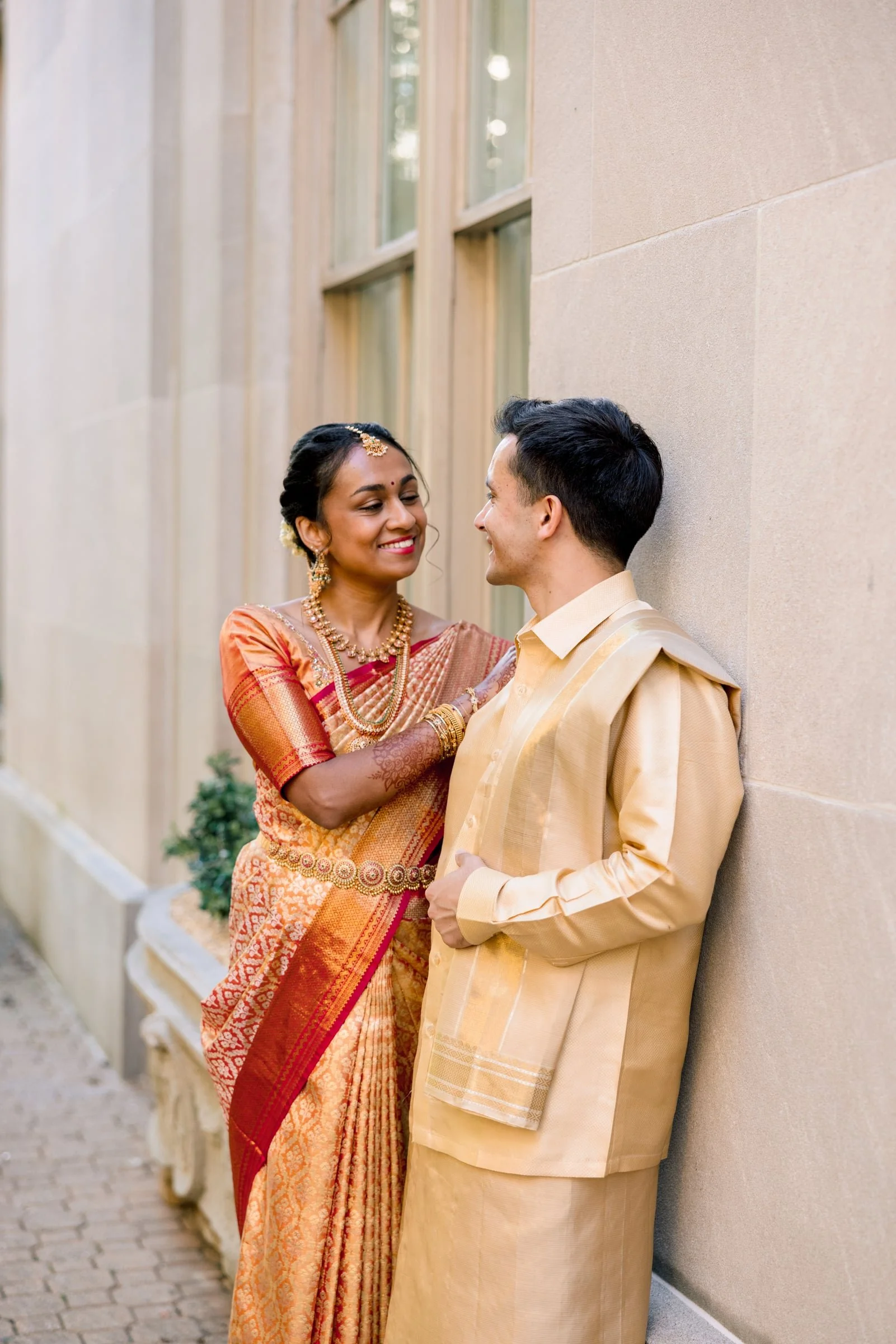 A couple dressed in traditional Indian attire, standing against a concrete wall outside a building, smiling at each other.