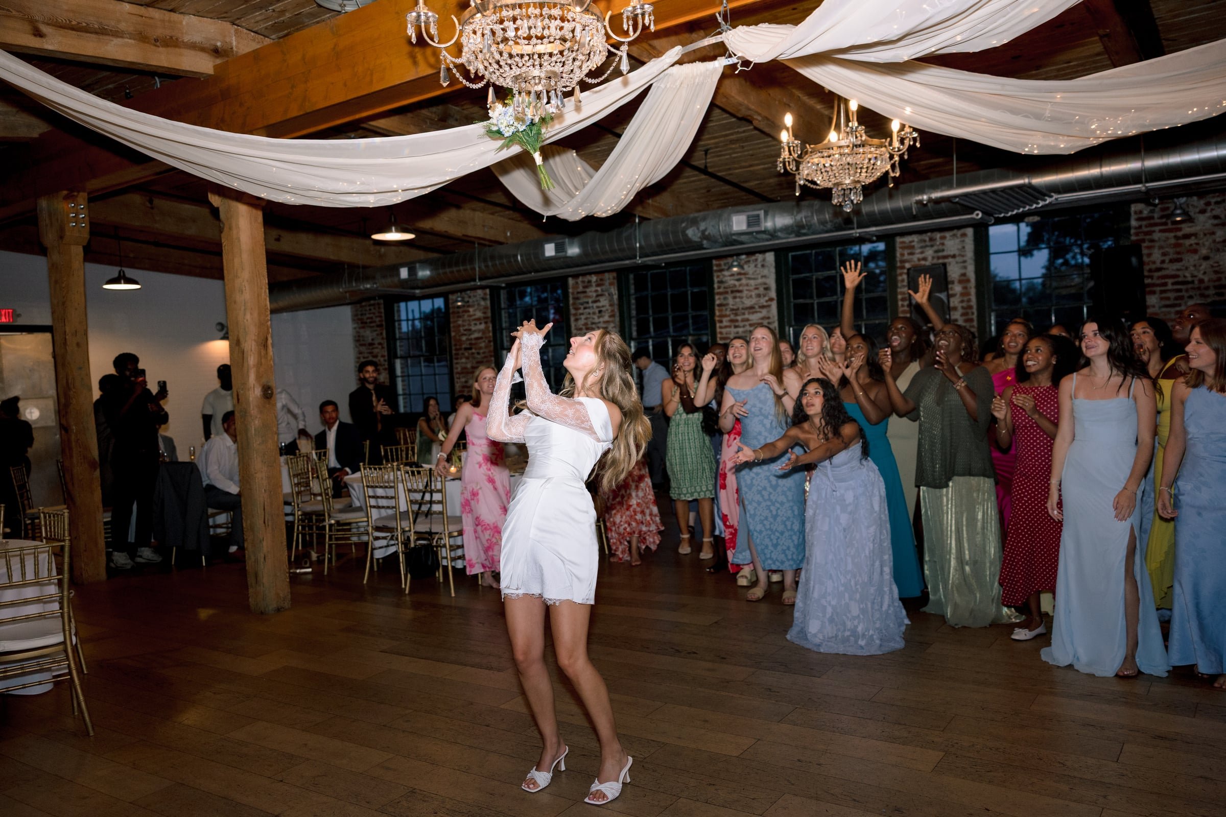 A group of women in colorful dresses watching a woman in a white dress dance at a wedding reception with chandeliers, draped fabric, and exposed brick walls.