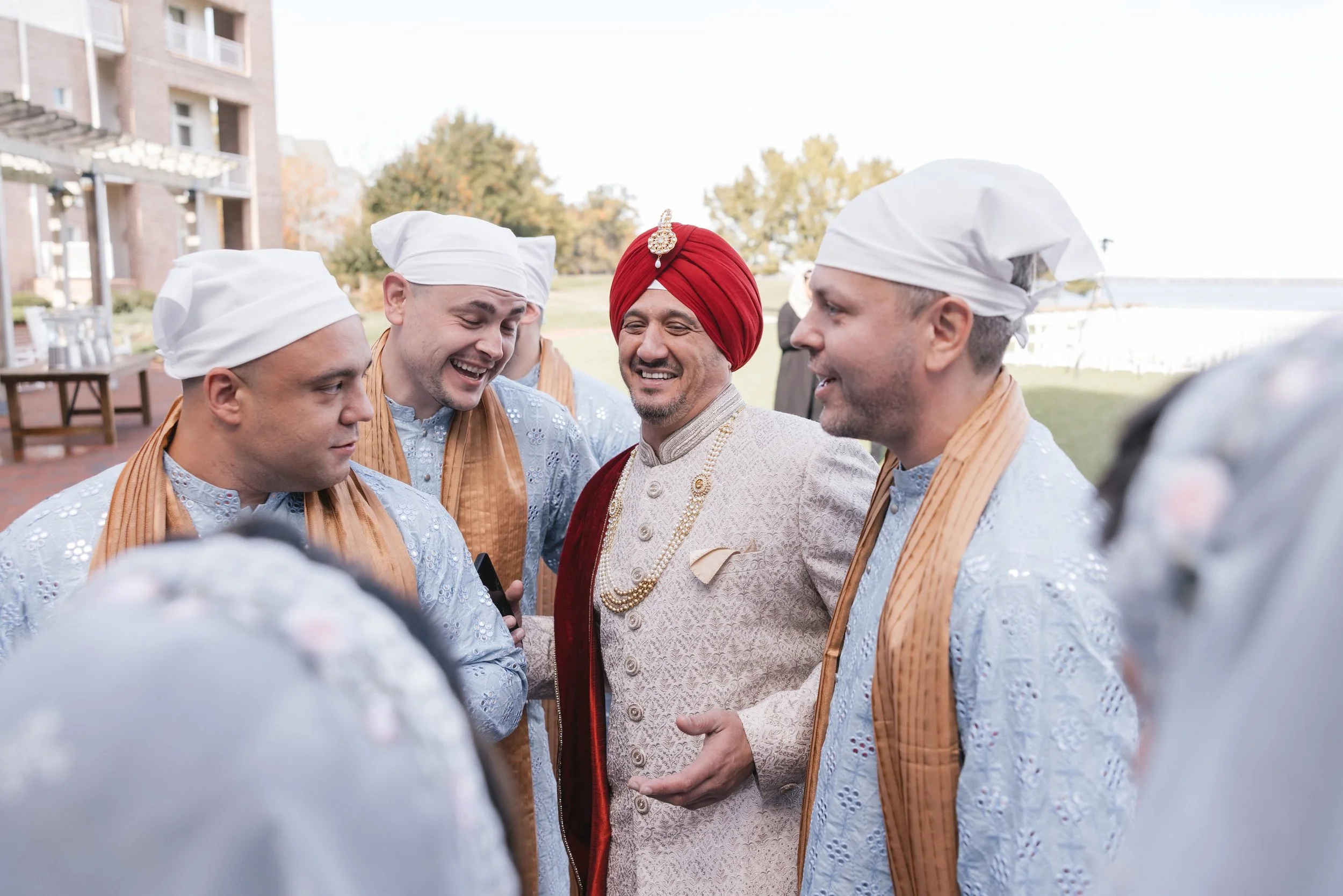 Group of men in traditional Indian attire, including turbans and embroidered jackets, gathered outdoors engaging in conversation, with a background of trees and a building.