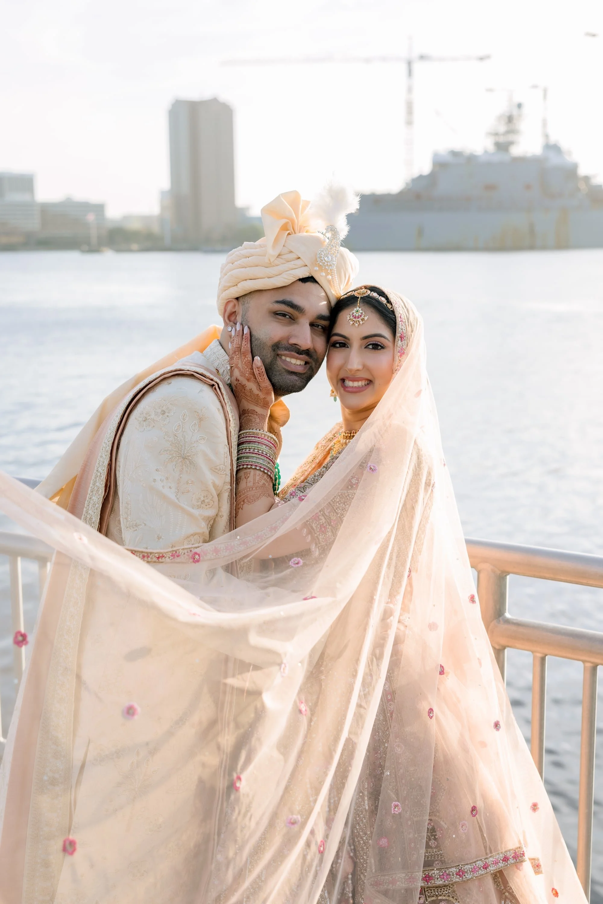 An interracial bride and groom dressed in traditional Indian wedding attire stand close together on a boat with water and city skyscrapers in the background. The groom wears a cream sherwani and turban with embroidery, and the bride wears a peach and