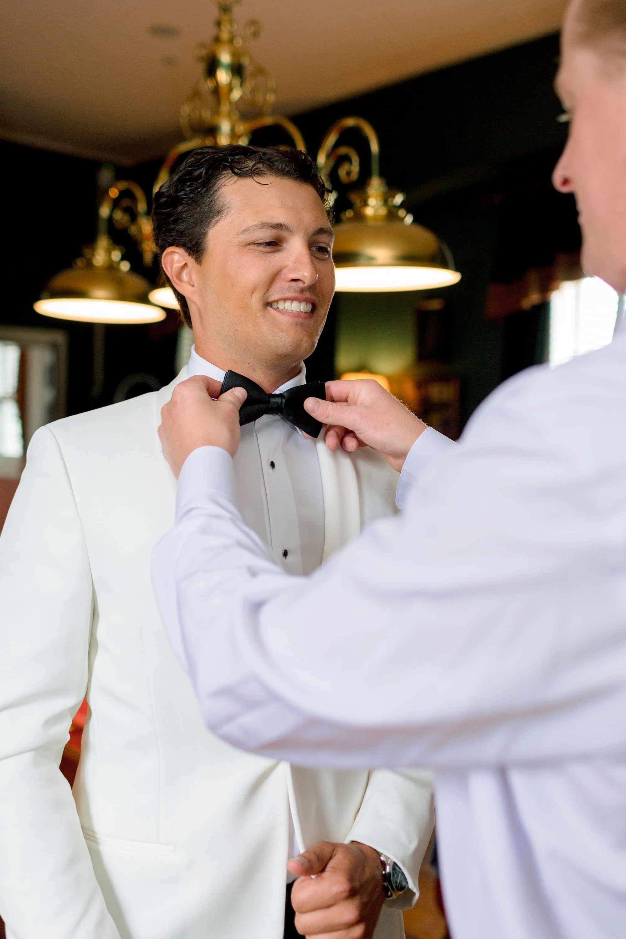 A man in a white tuxedo is being assisted with a black bow tie by another person. The man is smiling and adjusting his bow tie while the other person assists. The background shows warm lighting and decorative fixtures.