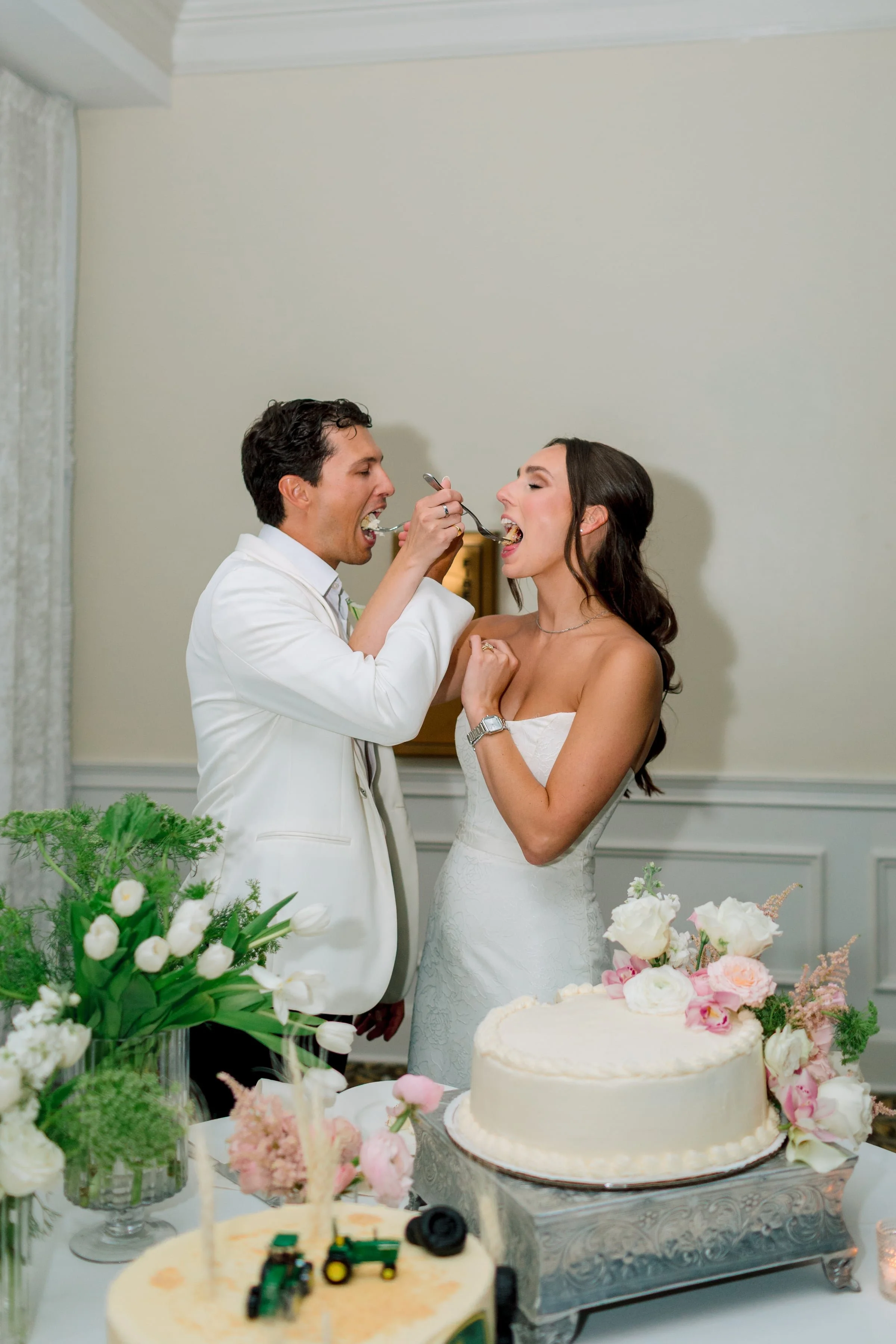 A bride and groom are feeding each other wedding cake at their reception with a decorated table of flowers and a wedding cake in the foreground.