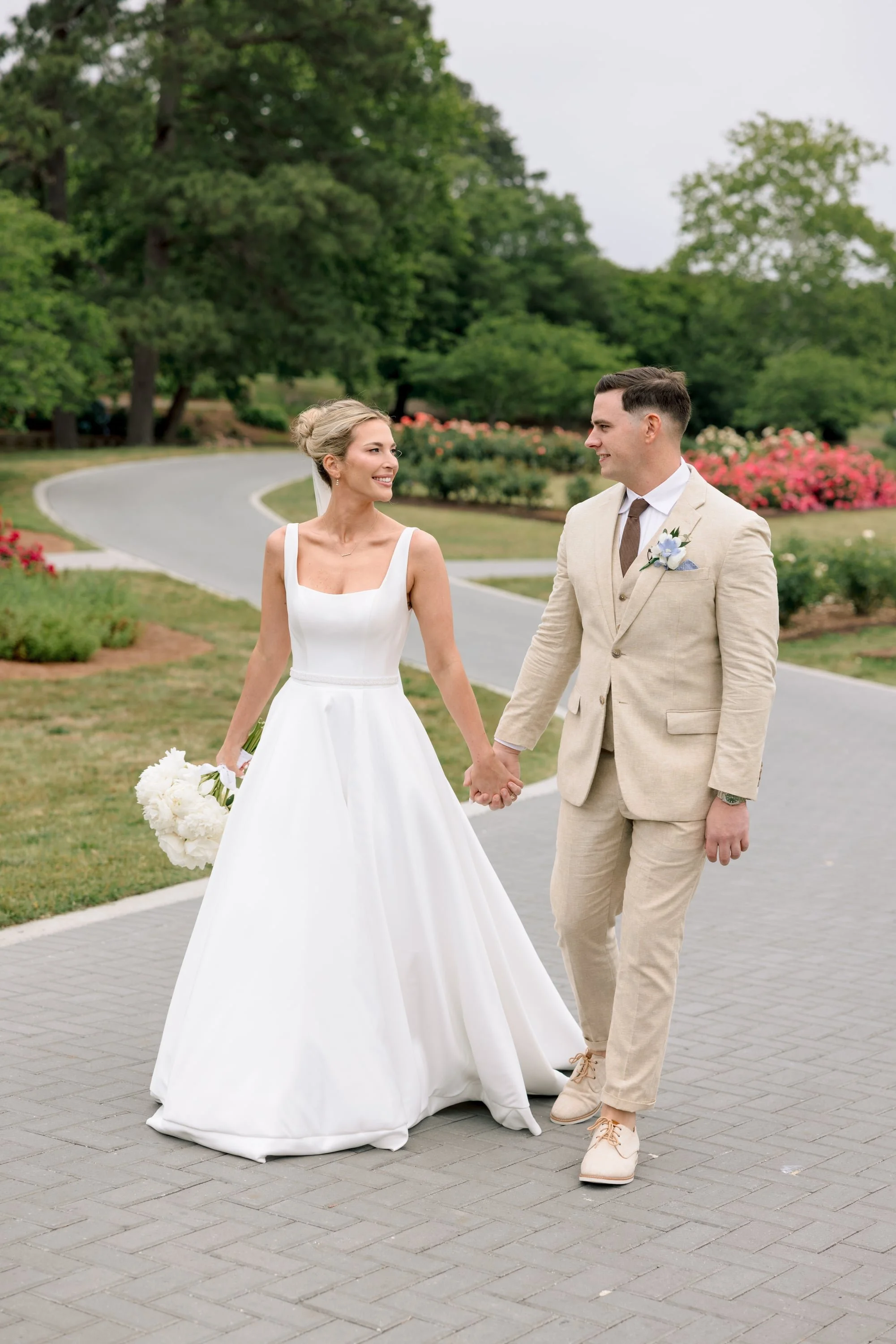 A bride and groom holding hands outdoors on a paved pathway, surrounded by lush green trees and colorful flowers, smiling at each other.