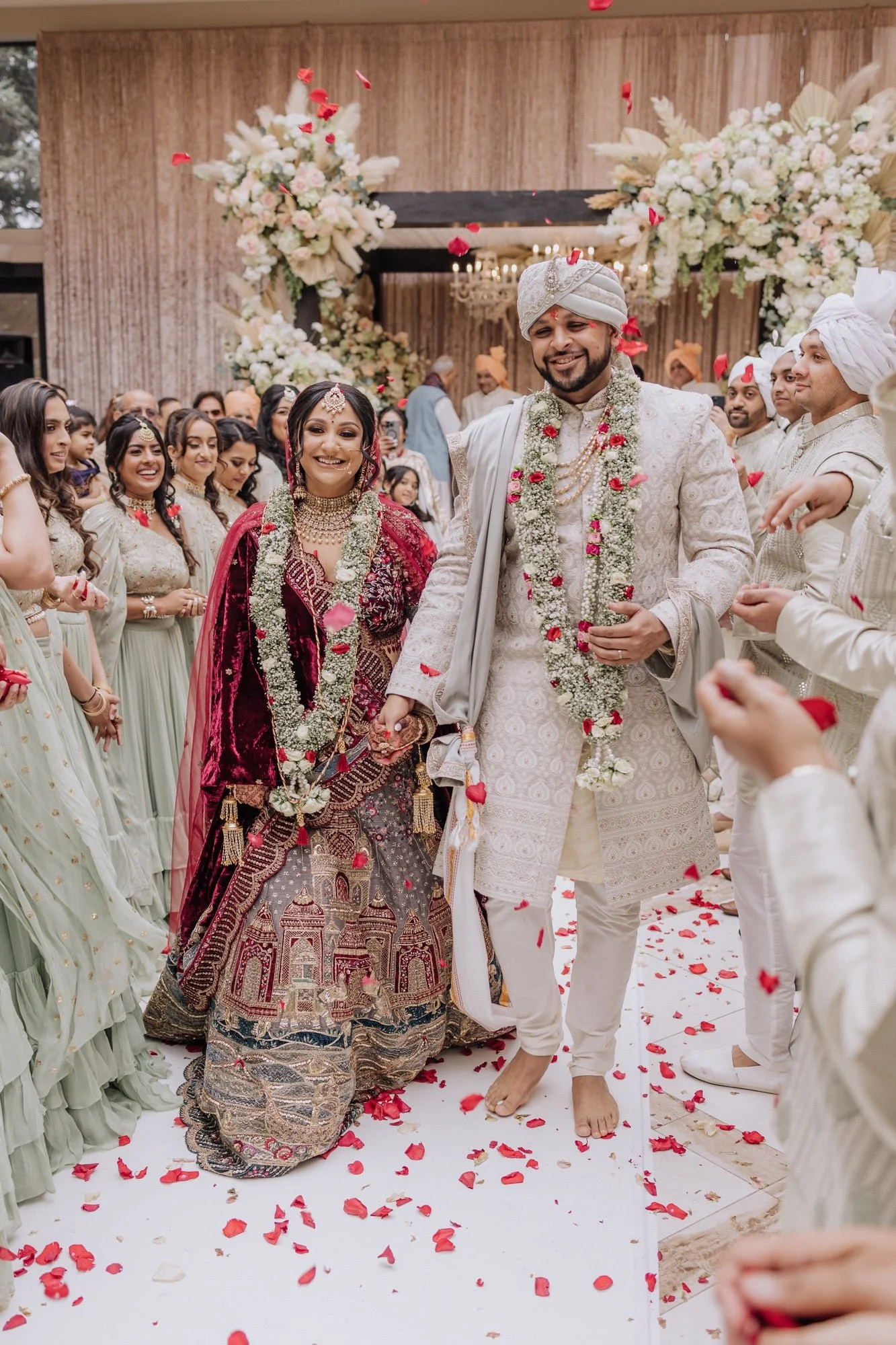 Indian bride and groom holding hands at their wedding ceremony, surrounded by family and friends, with flower petals on the floor and floral arrangements in the background.