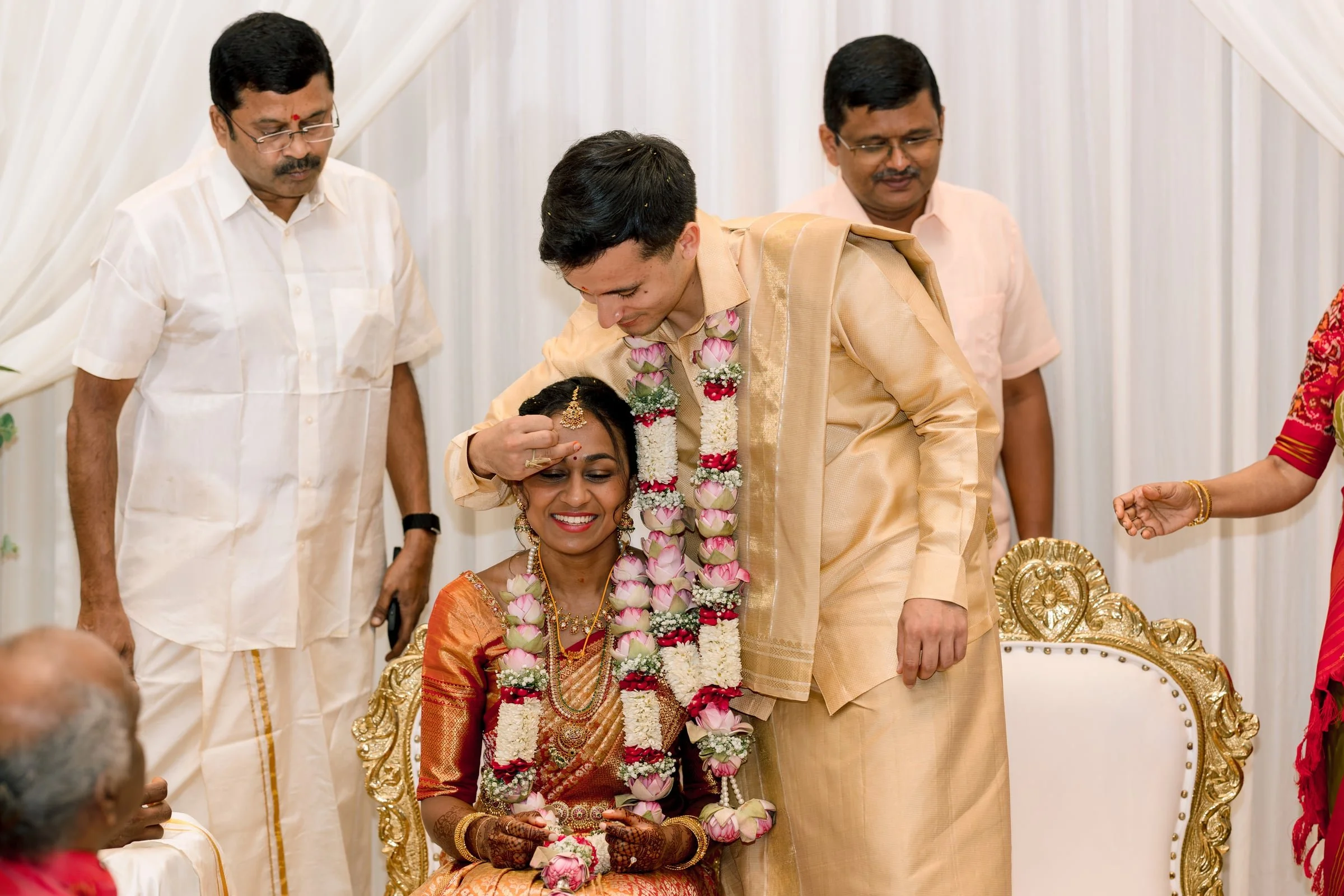 A traditional Indian wedding ceremony with a bride and groom dressed in ornate attire, surrounded by family members, in a decorated indoor setting.