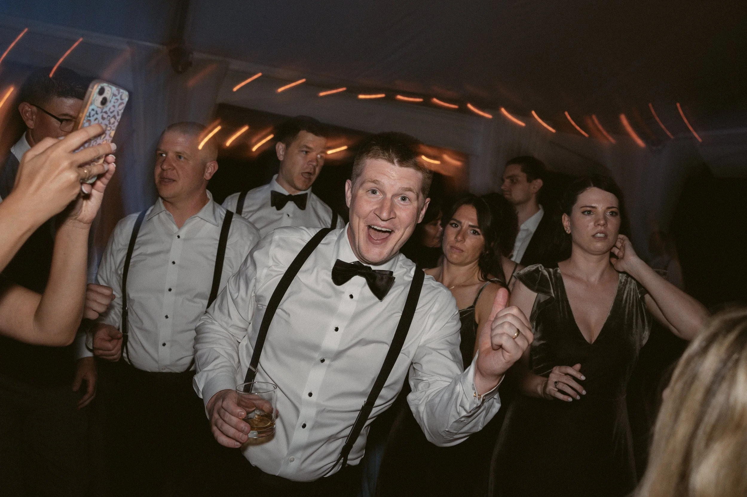 People dressed in formal attire at a party, with a man in a white shirt, black bow tie, and suspenders smiling and holding a drink, while others are talking or taking photos in a dimly lit room with orange string lights on the ceiling.