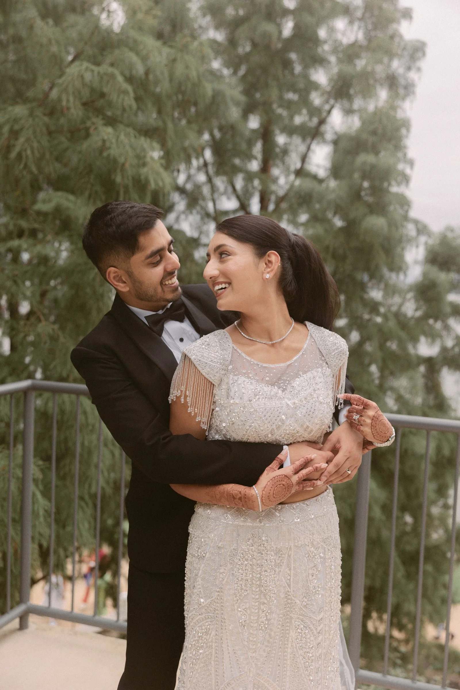 A happy couple in wedding attire embracing outdoors with trees in the background.