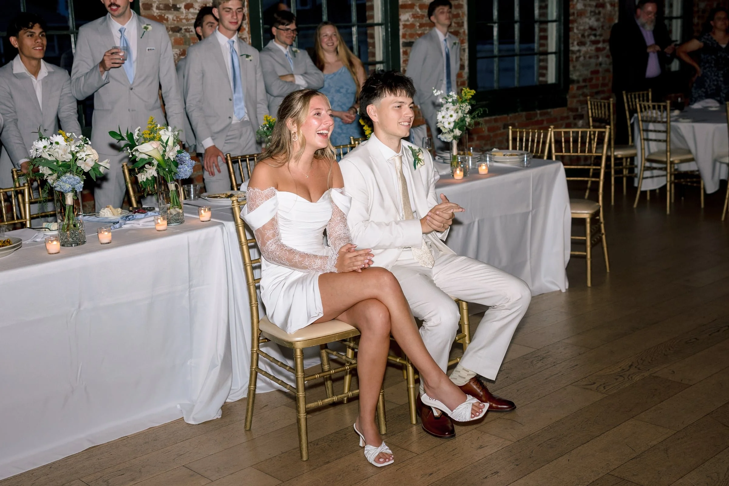 A wedding reception scene with a bride and groom sitting side by side at a table, smiling and laughing. The table is decorated with floral arrangements and candles. In the background, a group of men in suits and women, some in formal attire, are stan