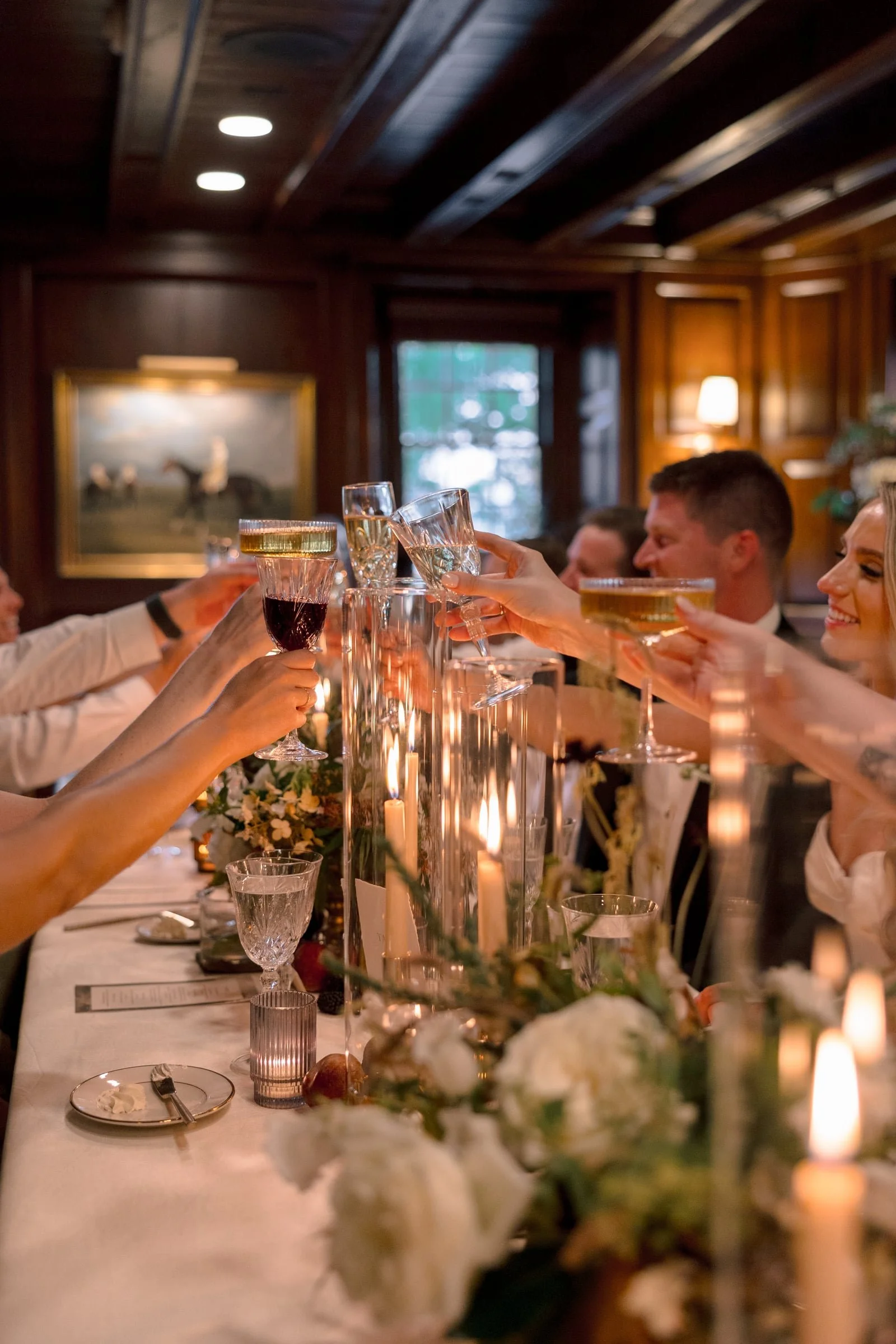 People raising glasses in a toast at a formal dinner with candlelit table decor and a wood-paneled room.